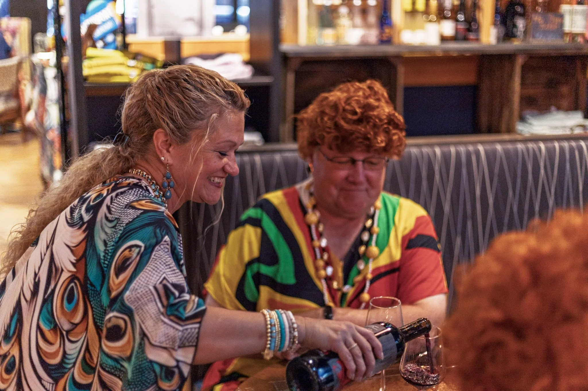 Two women sitting at a table pouring wine, smiling and enjoying, in a cozy indoor setting with shelves in the background.