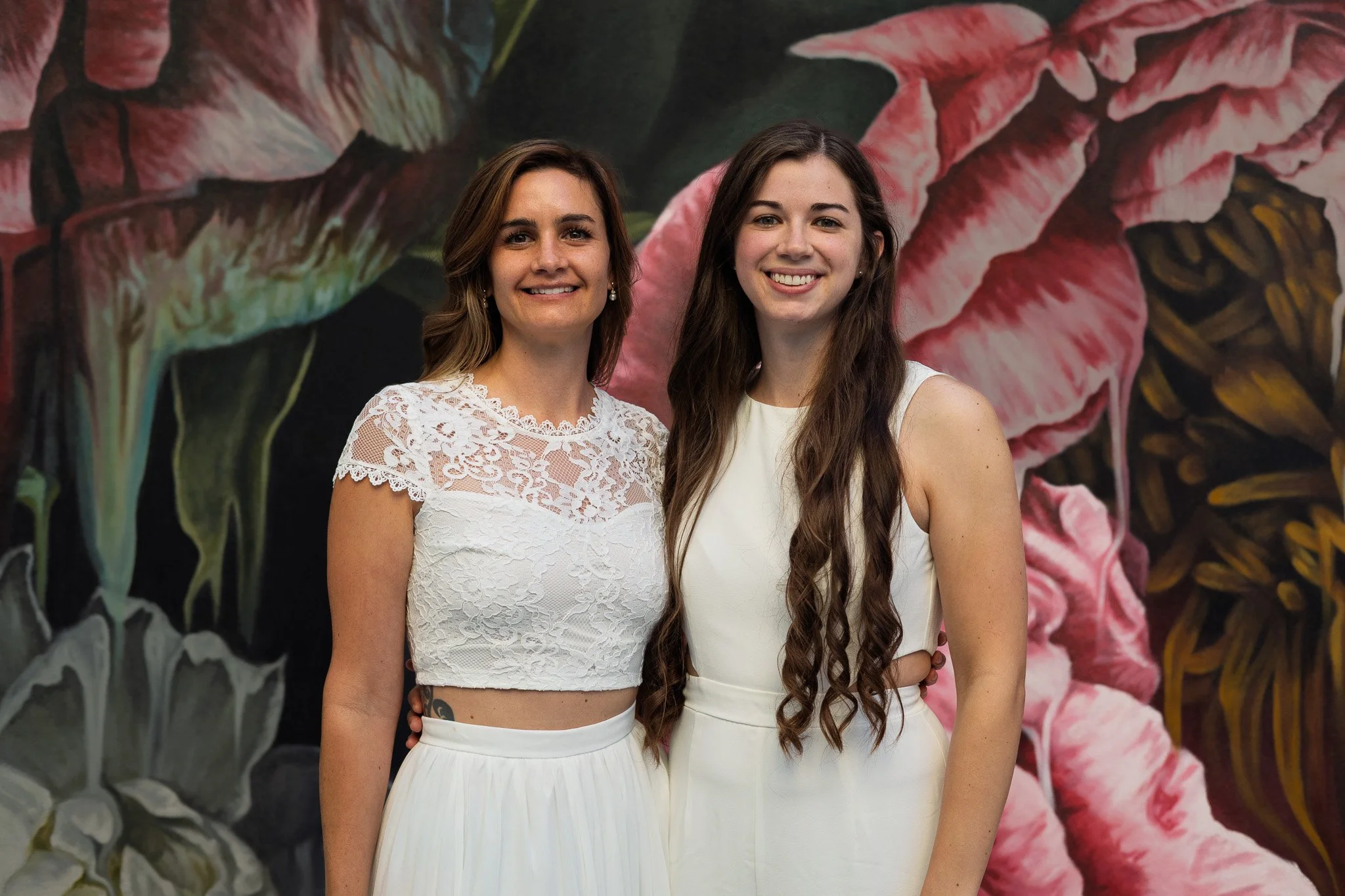 Two young women in white dresses standing close together in front of a large floral mural, smiling at the camera.