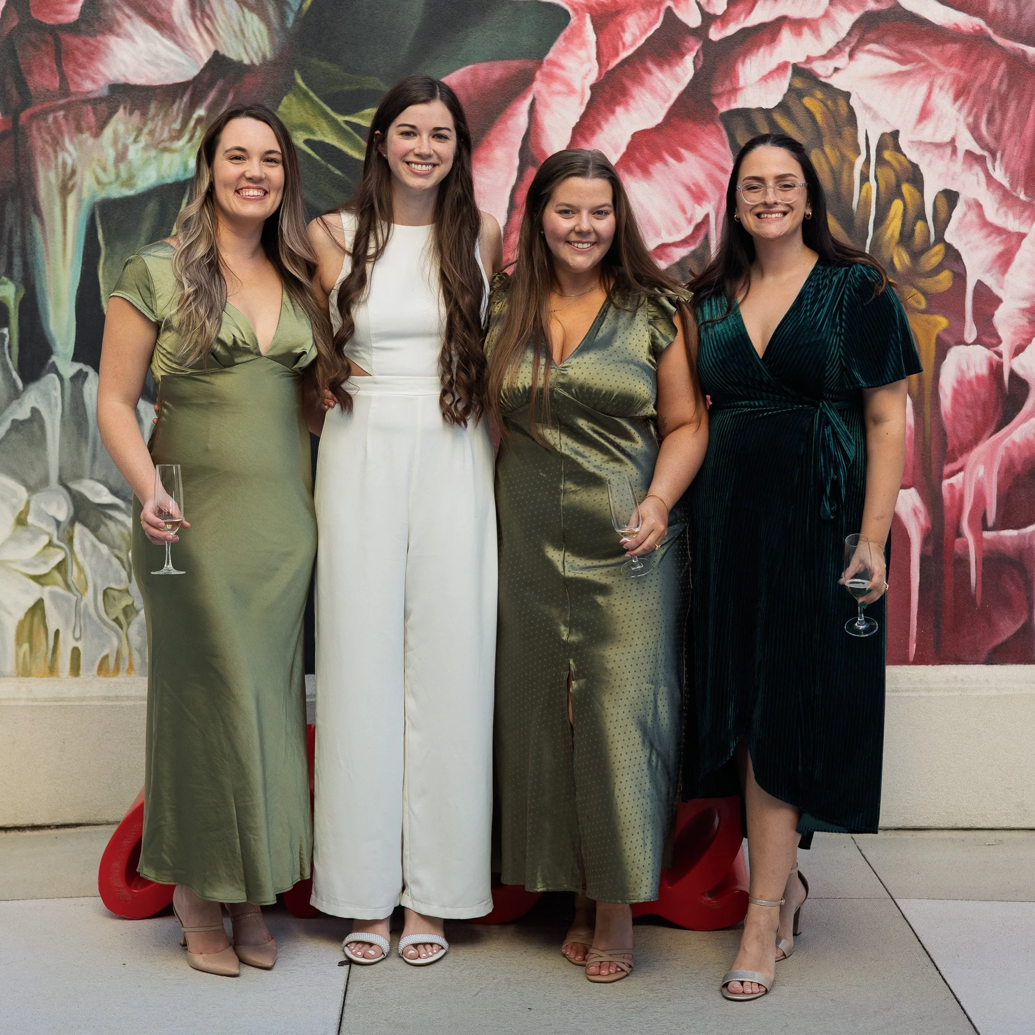 Four women dressed in elegant dresses standing together, smiling, holding champagne glasses, in front of a large floral mural.