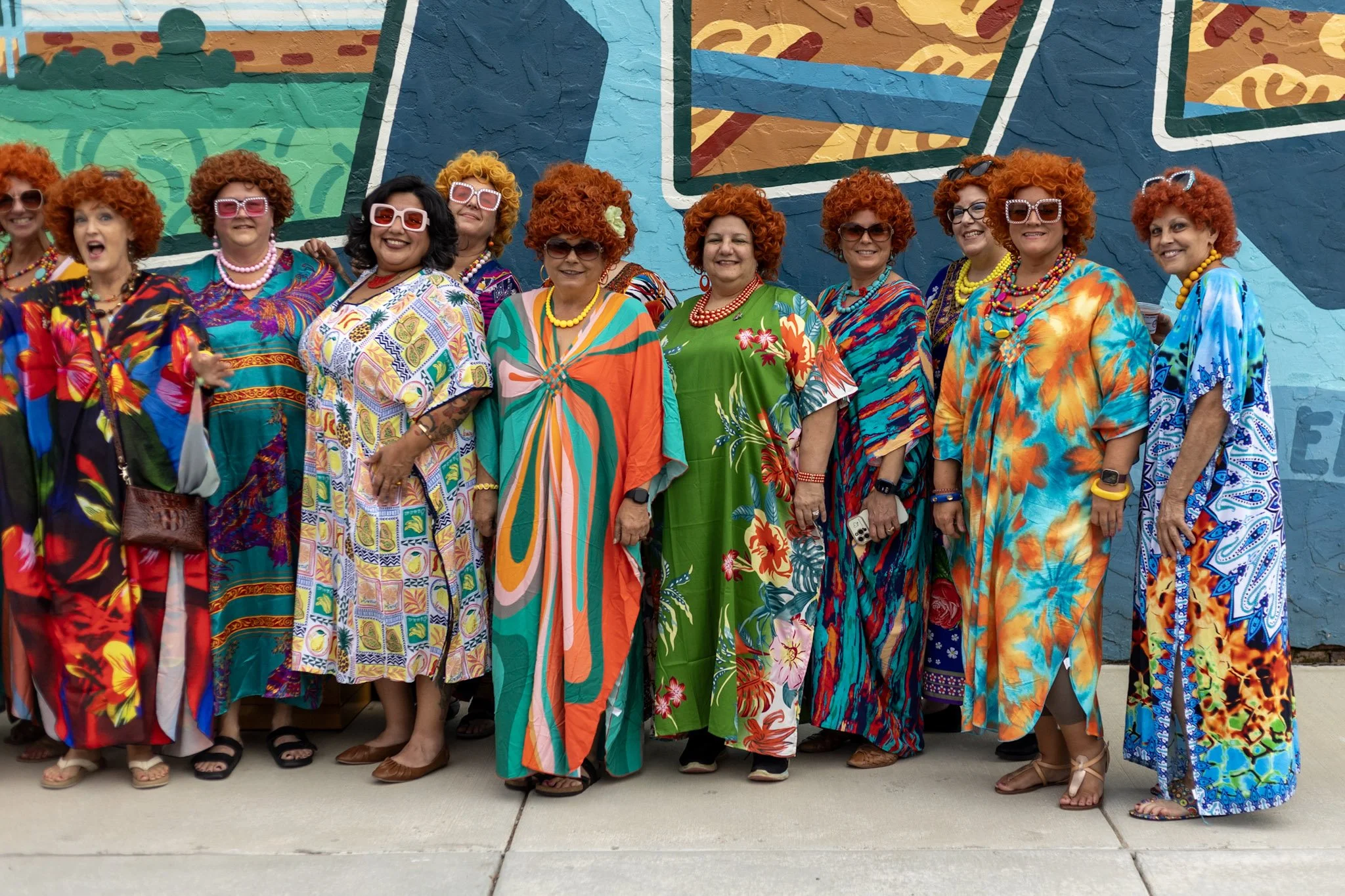 A group of women dressed in colorful, floral-patterned dresses and wearing sunglasses, standing in front of a mural.