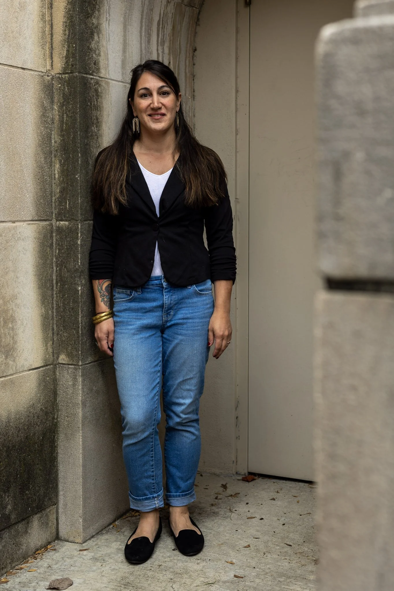 A woman standing in a narrow outdoor alleyway with stone walls and a doorway, smiling at the camera.
