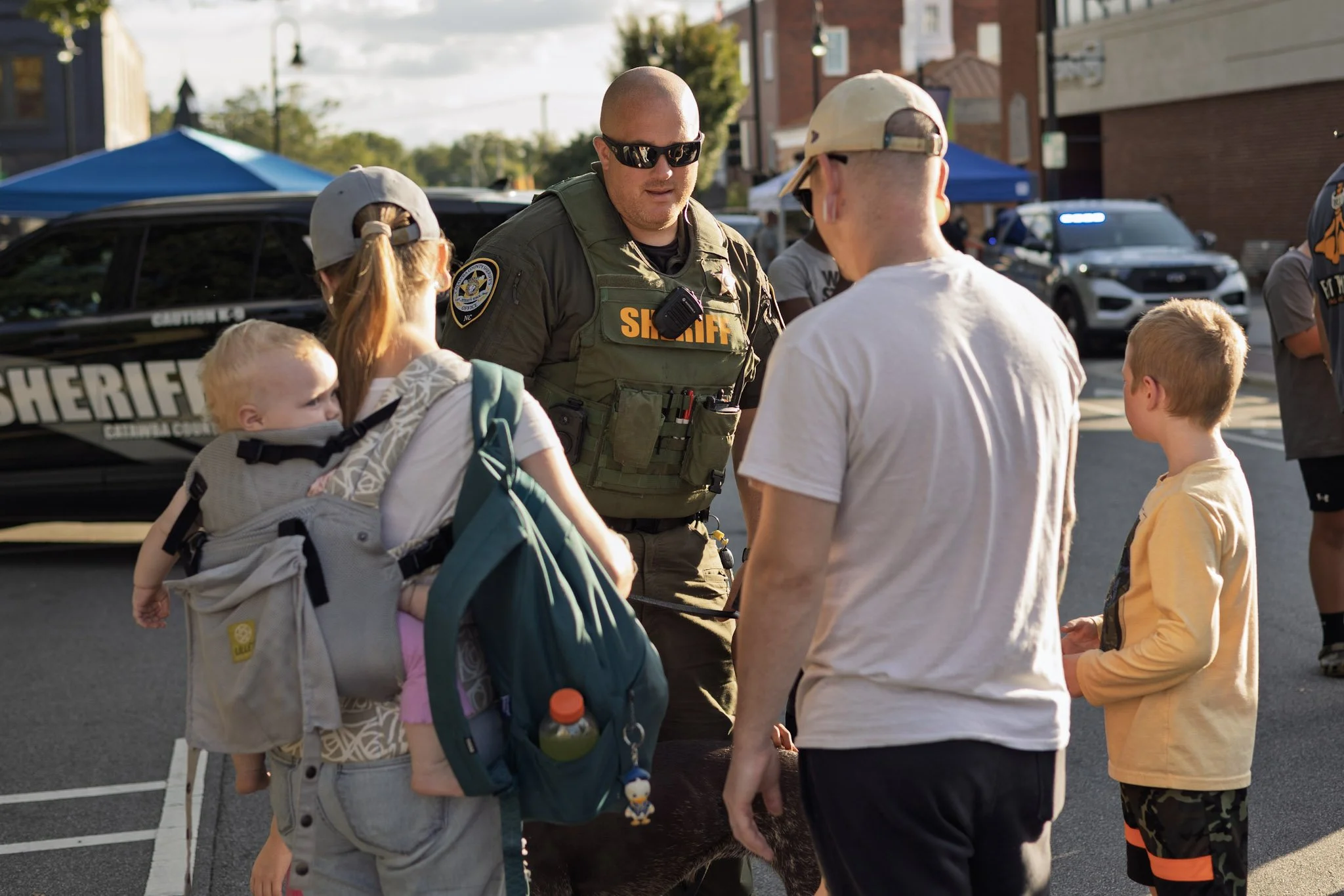 A police officer in uniform talking to a family and children during an outdoor event with police cars in the background.