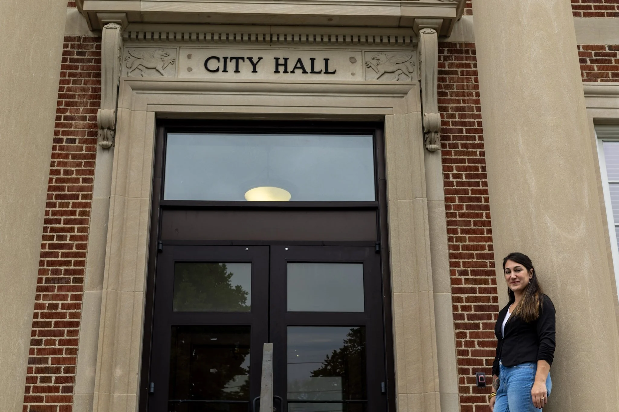 A young woman with long brown hair standing outside the entrance to a city hall building with a brick facade and large columns. The sign above the door reads 'CITY HALL'.