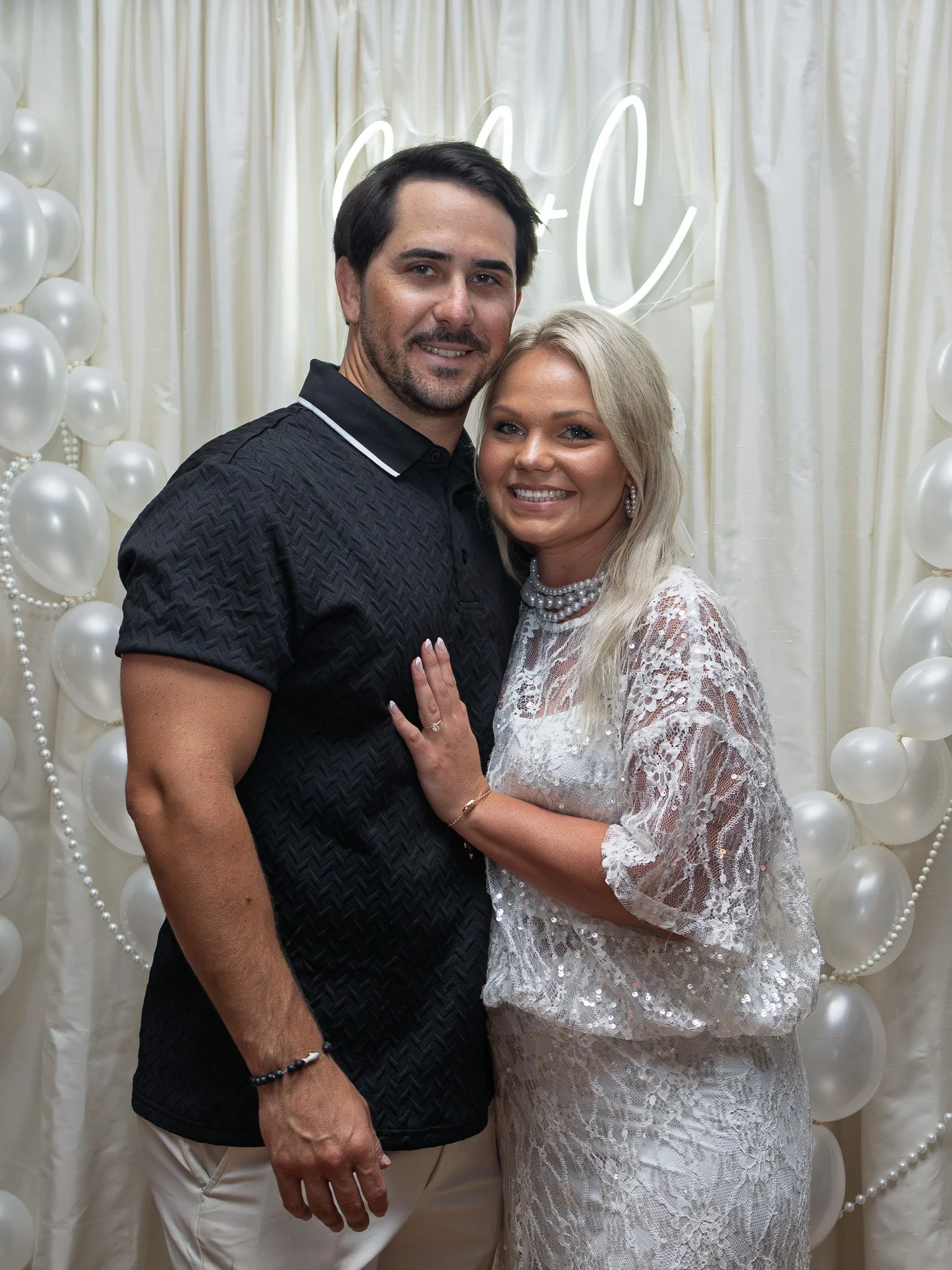Smiling man and woman standing close together, holding hands, in front of a white balloon backdrop and a neon sign, at a celebration or party.
