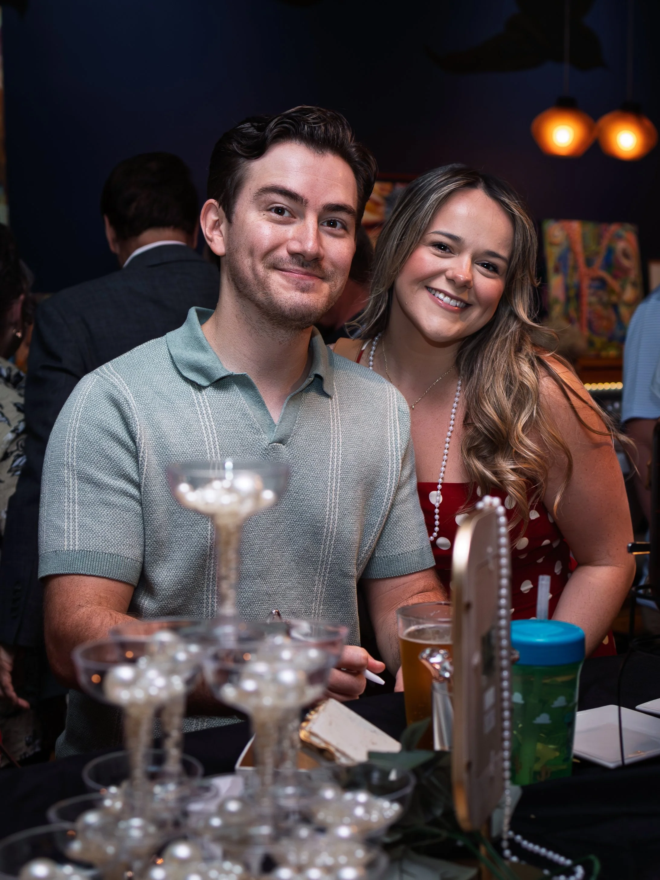 A smiling man and woman standing at a social gathering with drinks and decorations in the background.