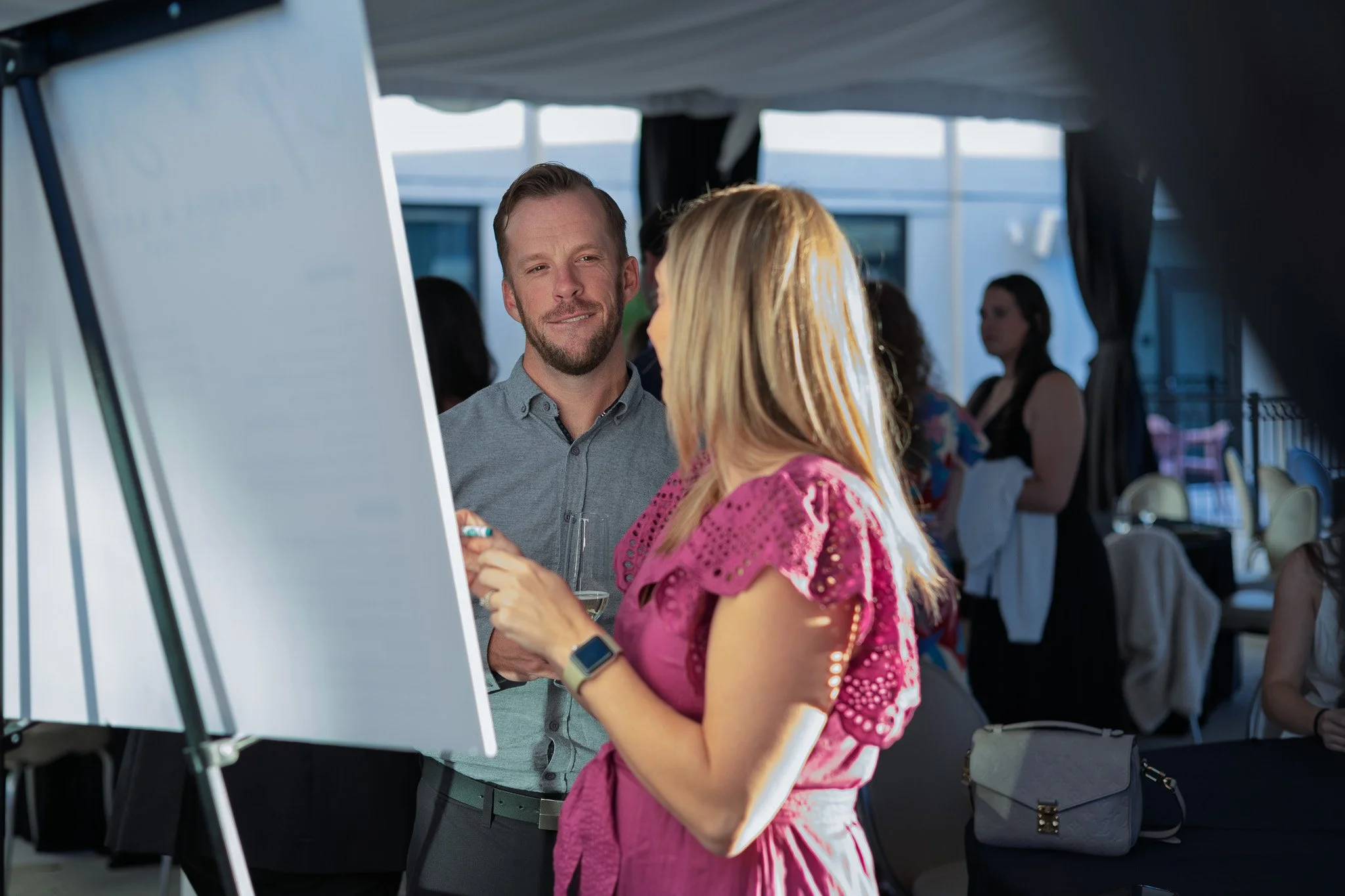 A man and woman are having a conversation at a social event, with the man holding a glass of wine, and a large whiteboard or presentation board in the foreground.