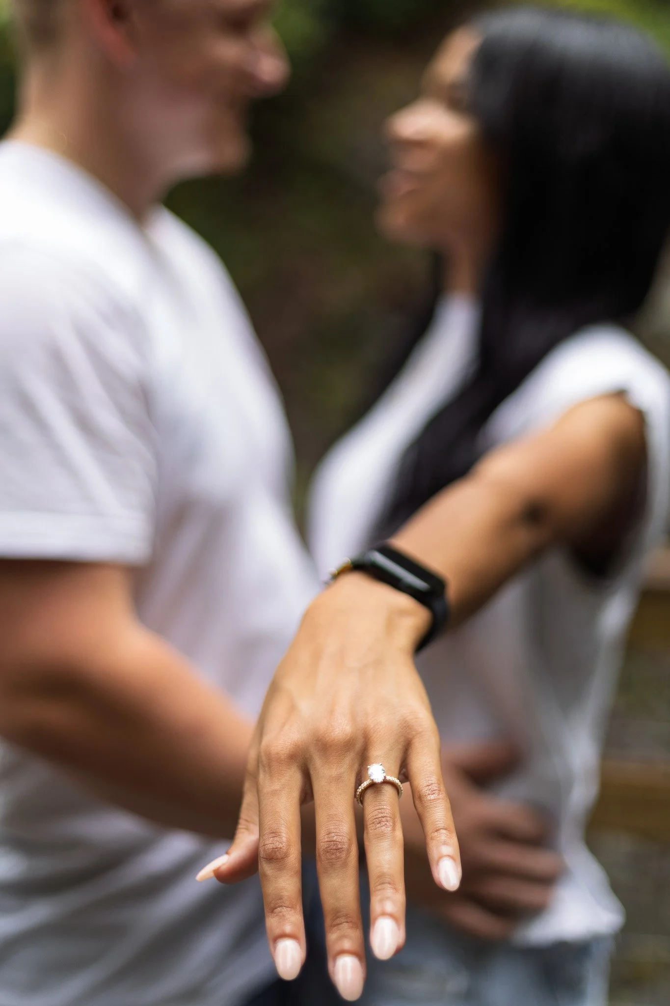 A woman shows off her engagement ring on her finger, with two people in the background smiling at each other.