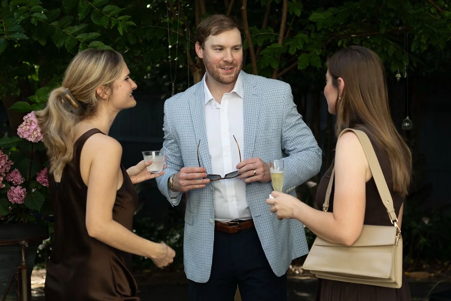 Three people, two women and one man, are having a conversation outdoors in a garden setting, each holding a drink, with the man in the center wearing a light-colored blazer and white shirt.