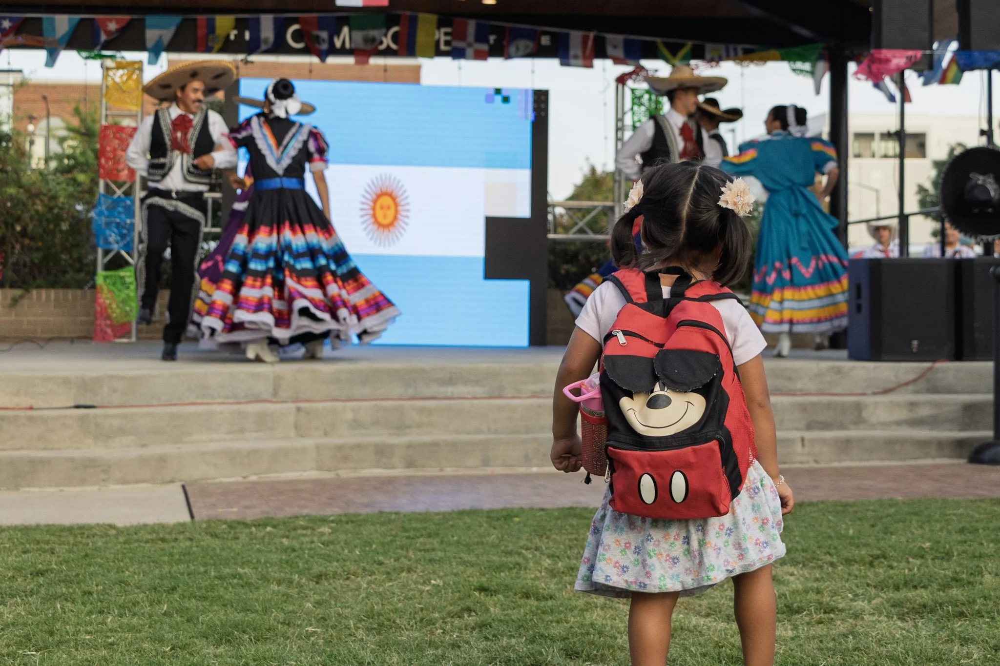 A young girl with a Mickey Mouse backpack watching a traditional Mexican dance performance on stage, with performers in colorful costumes and sombreros.