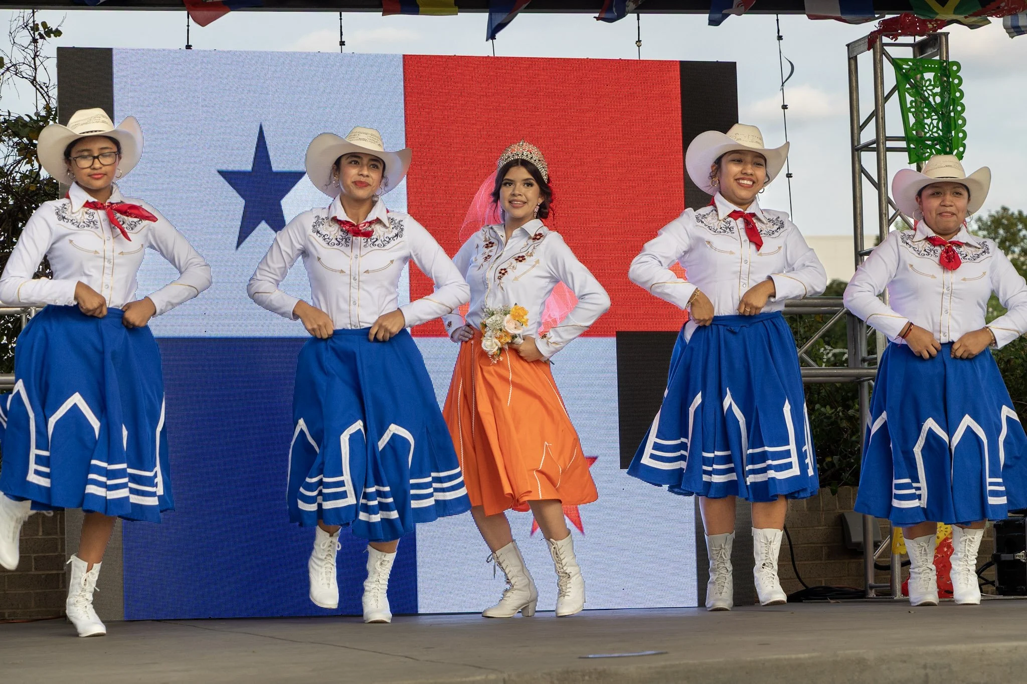 Five women in traditional Mexican folk dance costumes stand on a stage, with a large flag in the background. Four women wear white shirts, blue skirts, white cowboy hats, and white boots. One woman in the middle wears an orange skirt, white shirt, an