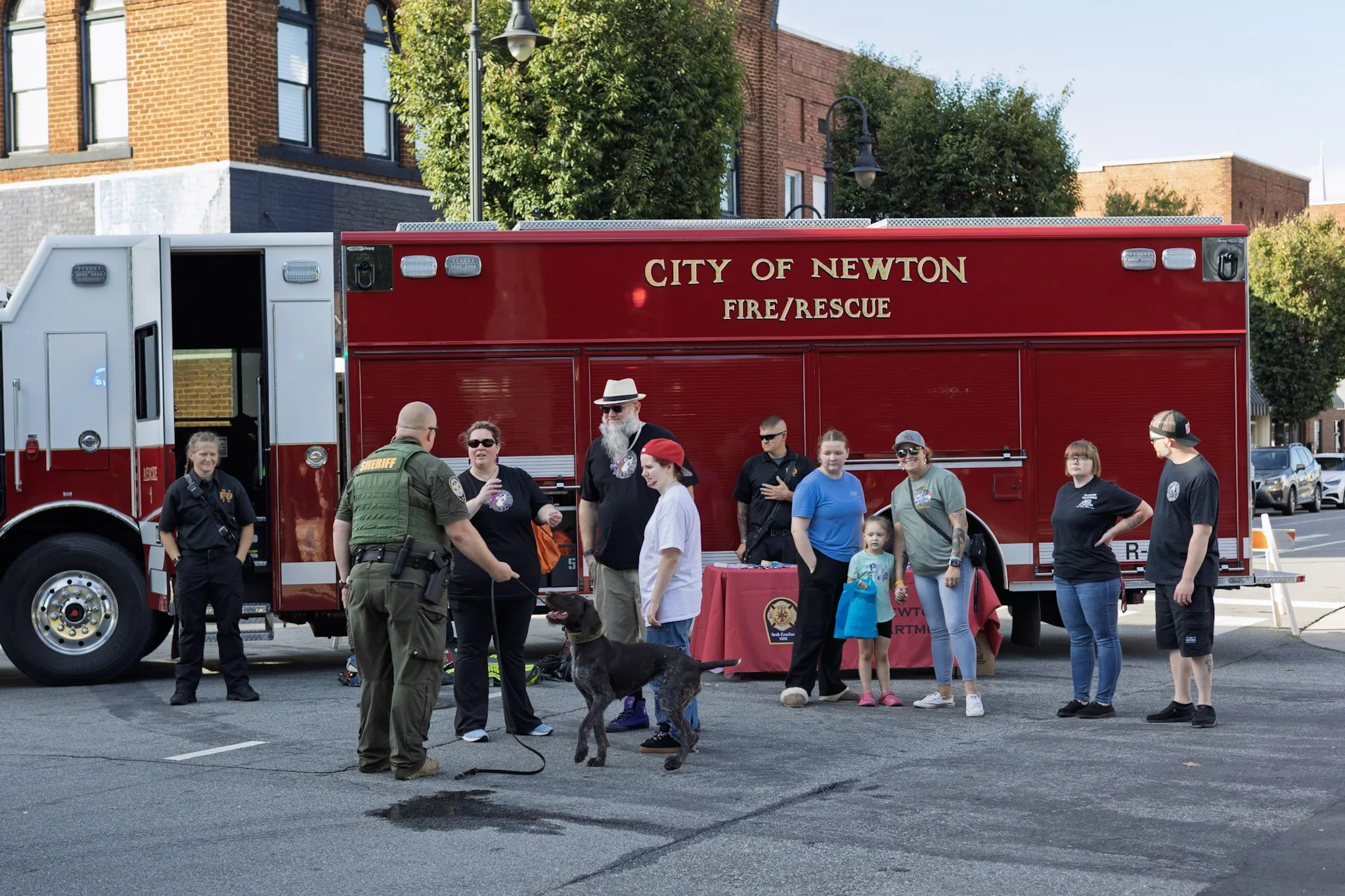 People gather in front of a red City of Newton Fire/Rescue truck for a community event. Firefighters and civilians, including kids, interact, and a dog is present. The event is outdoors in a city street with brick buildings and trees.