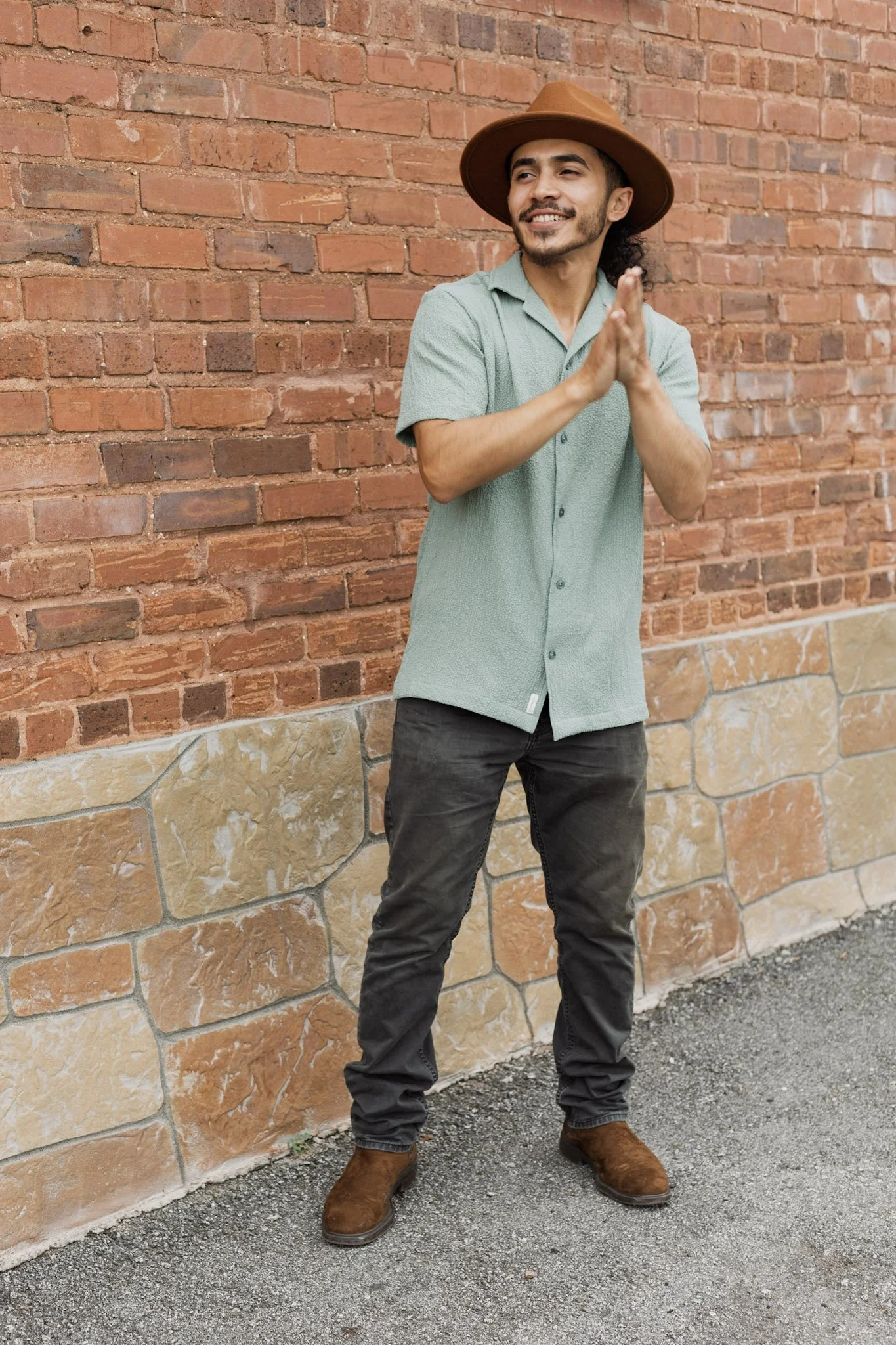 A man with a beard and mustache, wearing a tan wide-brimmed hat, green short-sleeved button-up shirt, black jeans, and brown boots, standing against a red brick wall with his hands together and smiling.