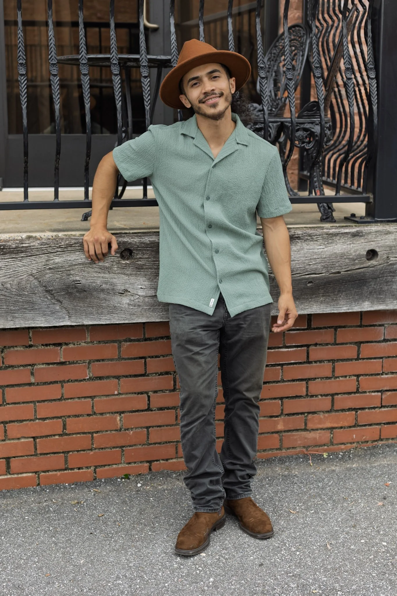 A young man with a beard and curly hair wearing a brown hat, green short-sleeve button-up shirt, black pants, and brown boots leaning against a brick wall outdoors.