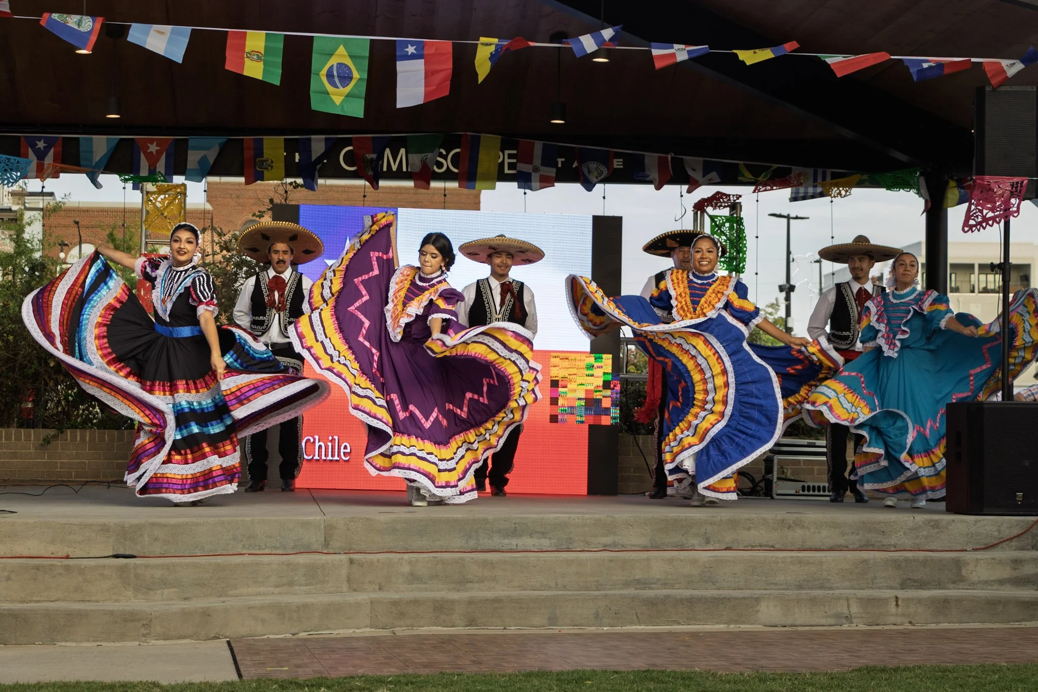 Group of dancers dressed in colorful traditional Mexican attire performing on an outdoor stage decorated with international flags at a cultural event.