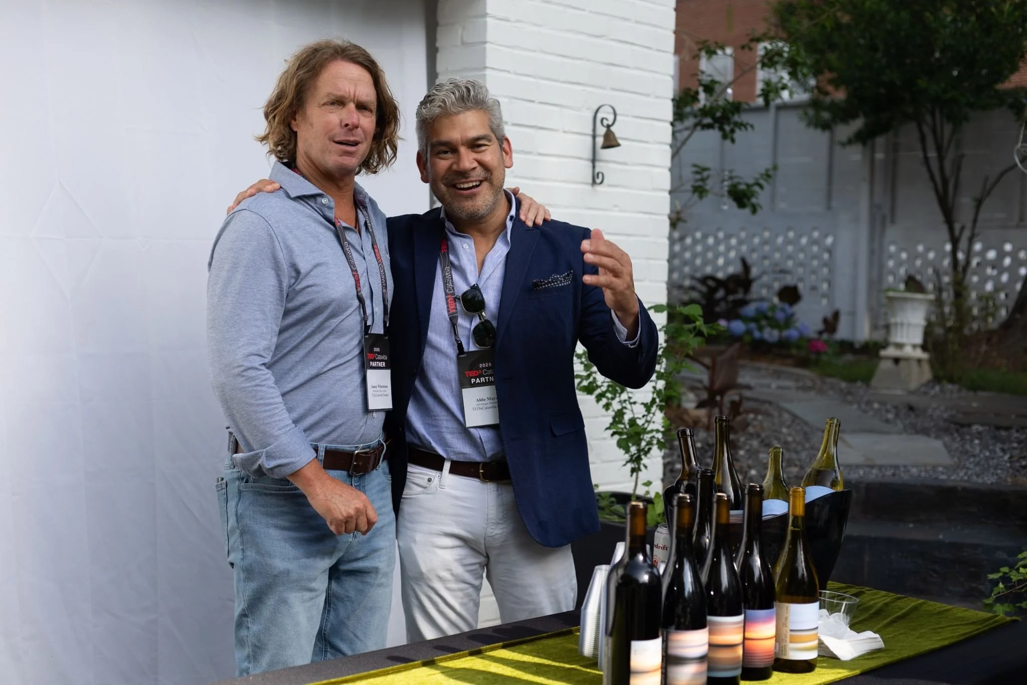 Two men standing close together with their arms around each other, smiling. They are outdoors at a table with multiple wine bottles, and they are wearing name badges around their necks.