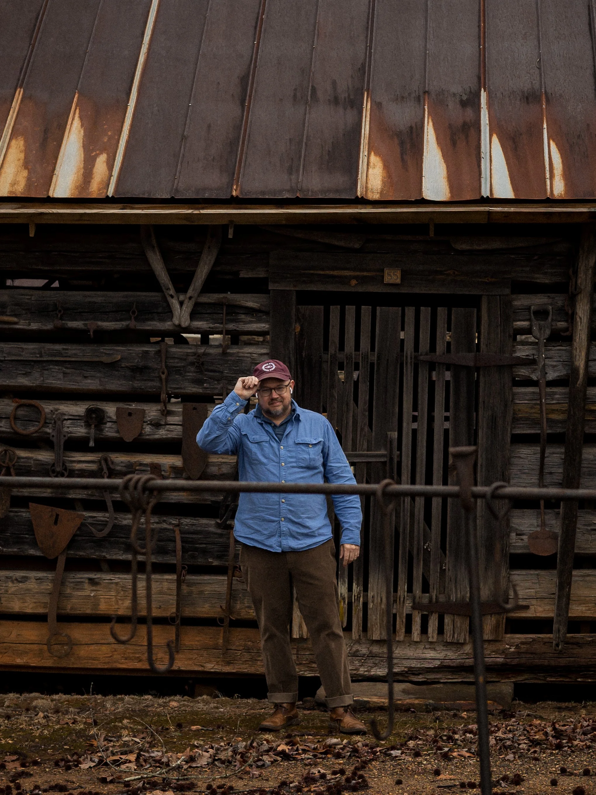 A man standing outdoors in front of a rustic wooden barn with tools hanging on the wall, wearing glasses, a blue shirt, brown pants, and a maroon cap, with autumn leaves on the ground.