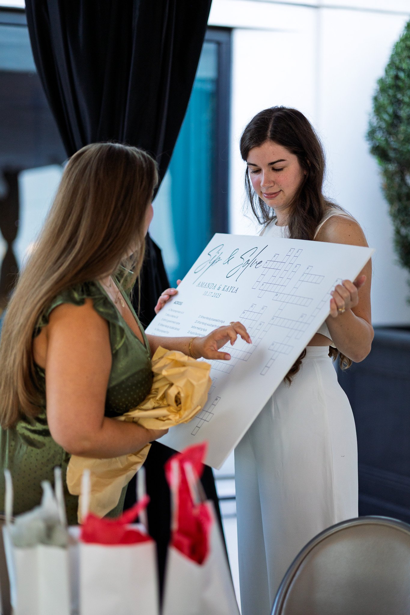 Two women at a wedding event, one in white dress exchanging a gift or card with another in a green dress with long blonde hair, holding a wrapped gift, in a decorated indoor setting.