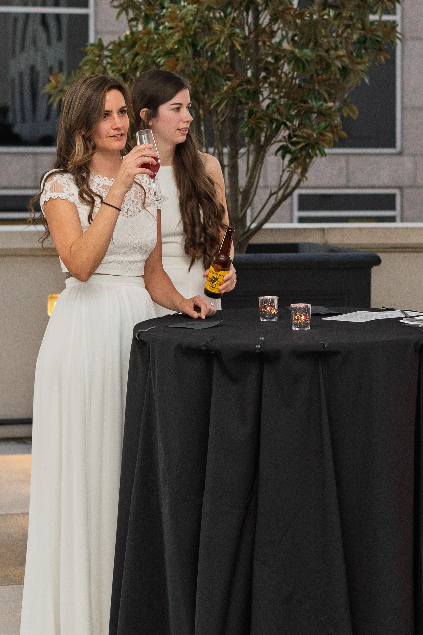 Two women in white dresses standing at a black table with candles during an outdoor event, with one woman holding a glass of wine and the other holding a beer bottle.