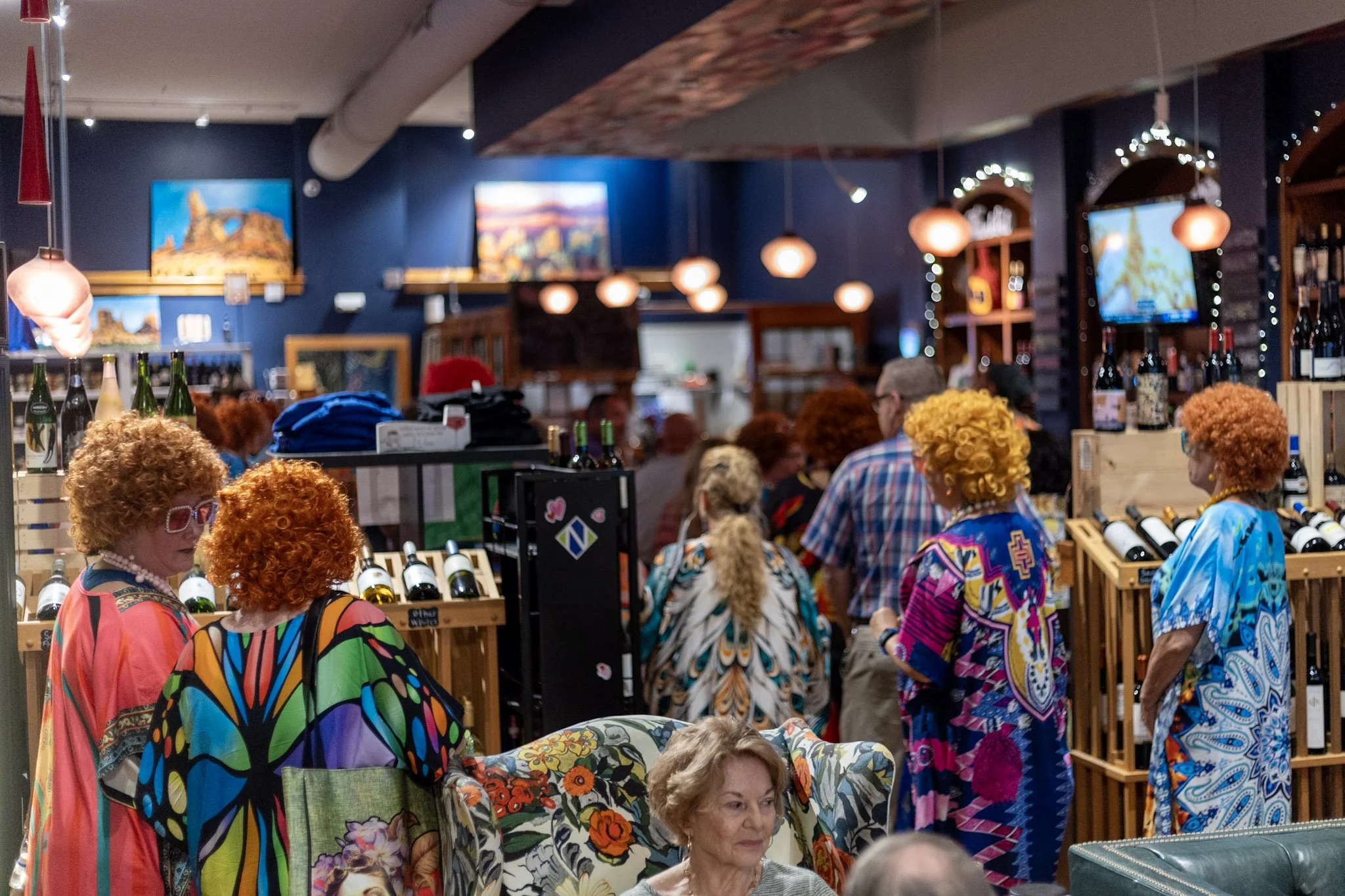 People dressed in colorful, patterned clothes and wigs, shopping for wine in a wine shop with blue walls, paintings, hanging lights, and shelves of wine bottles.