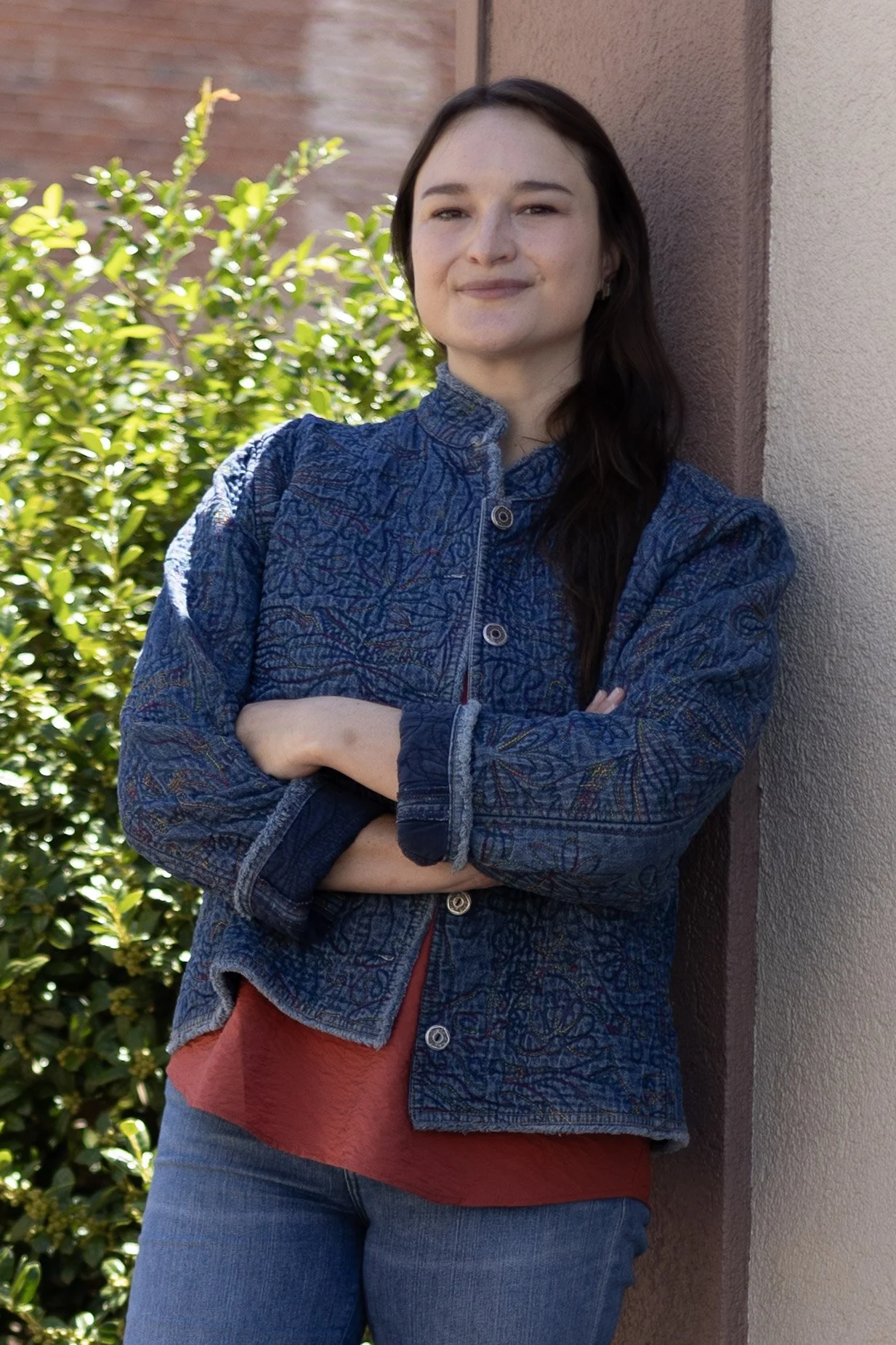 A woman with long dark hair standing outside near a wall, crossing her arms and smiling softly, with green bushes in the background.
