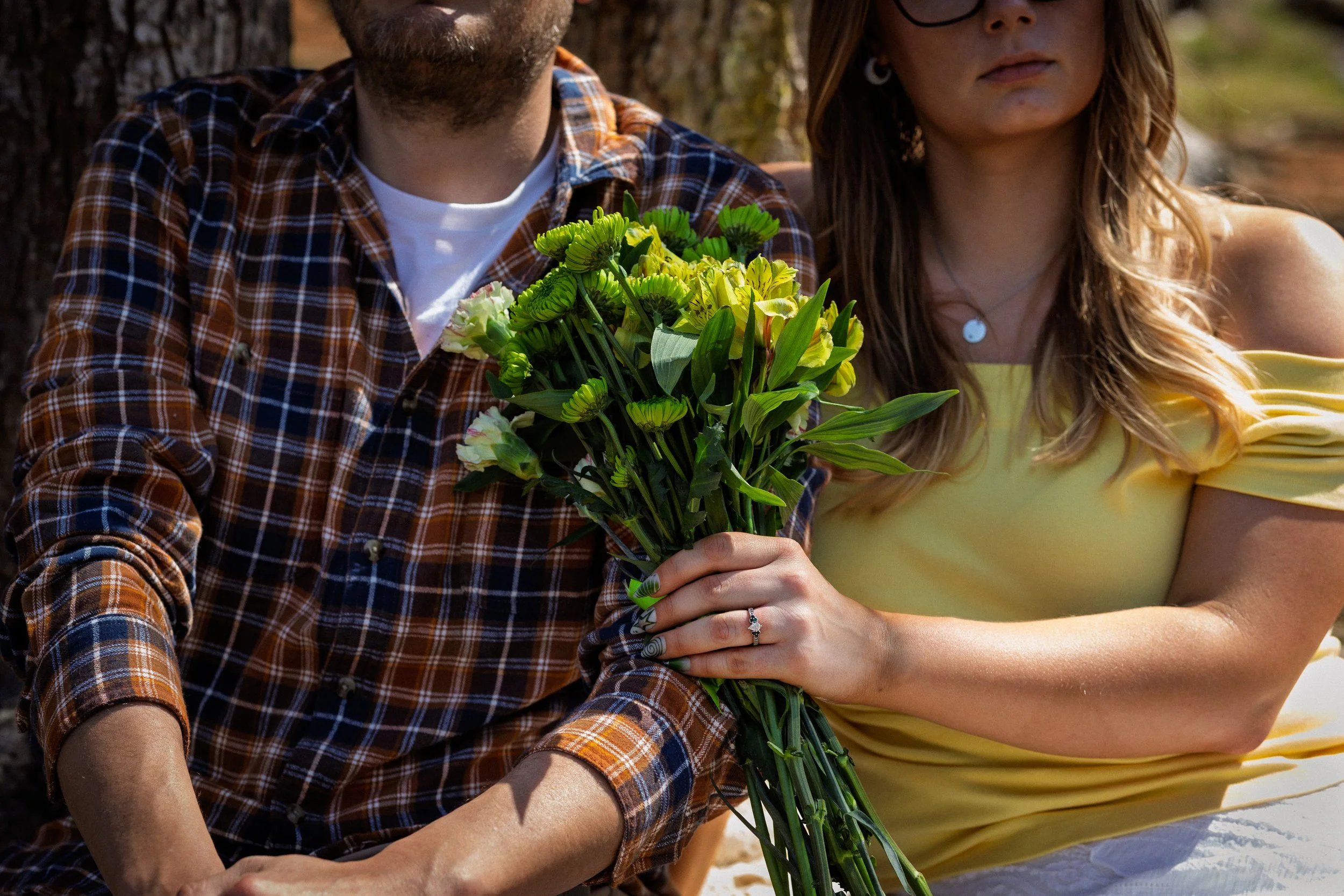 A man and woman sitting outdoors, holding a large bouquet of mixed green and yellow flowers, with trees in the background.