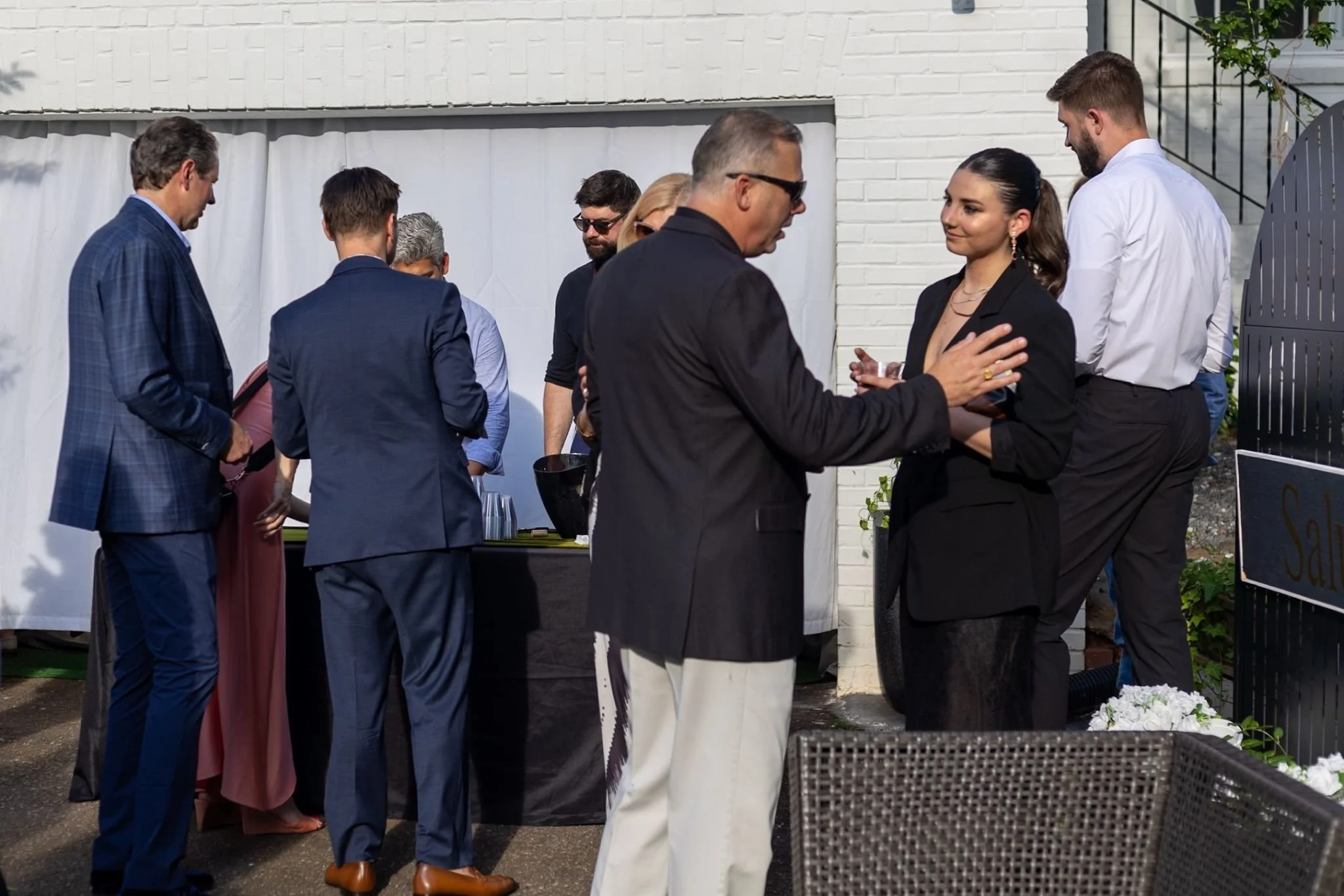 People gathered at an outdoor event, casually chatting by a white brick wall and a table with drinks.