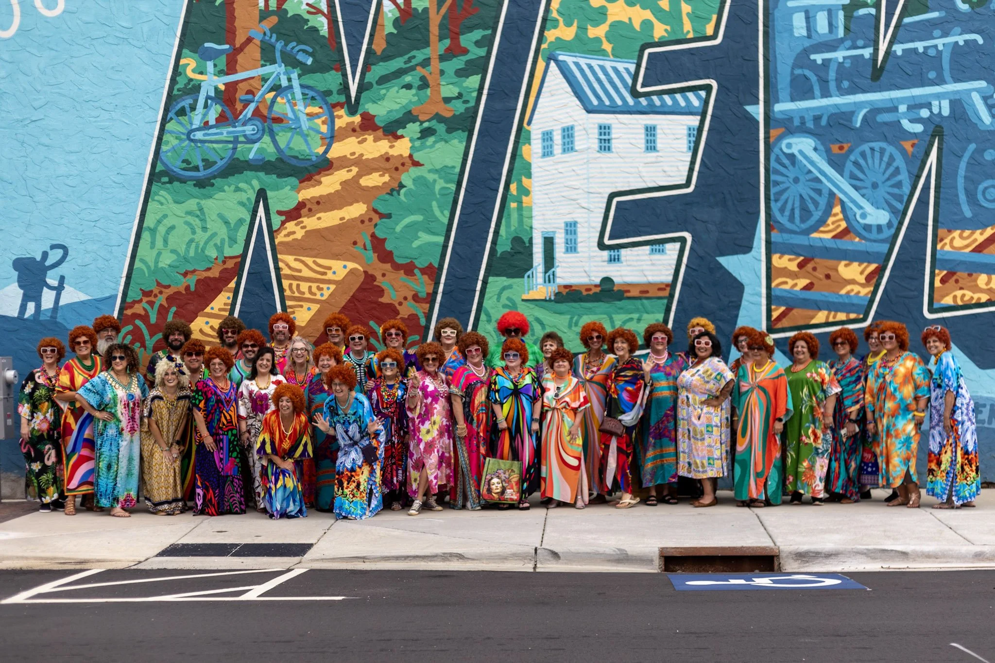 A group of women dressed in colorful, retro-style dresses with matching red wigs, posing in front of a vibrant mural on a city wall.