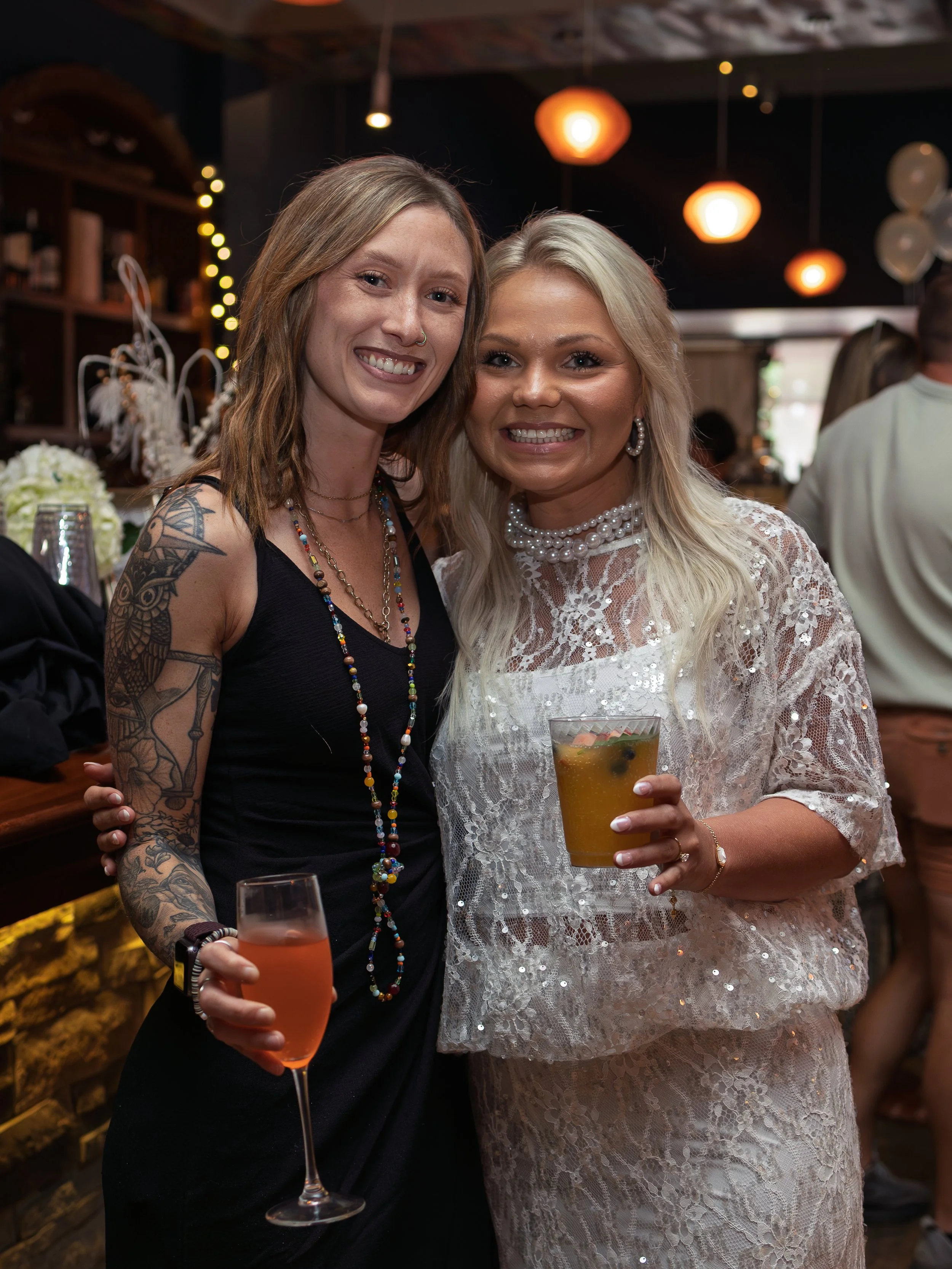 Two smiling women at a social gathering, holding drinks. One has tattoos on her arm and is wearing a black dress with colorful beaded necklaces. The other is dressed in a white lace dress with pearl jewelry. The background shows warm hanging lights a