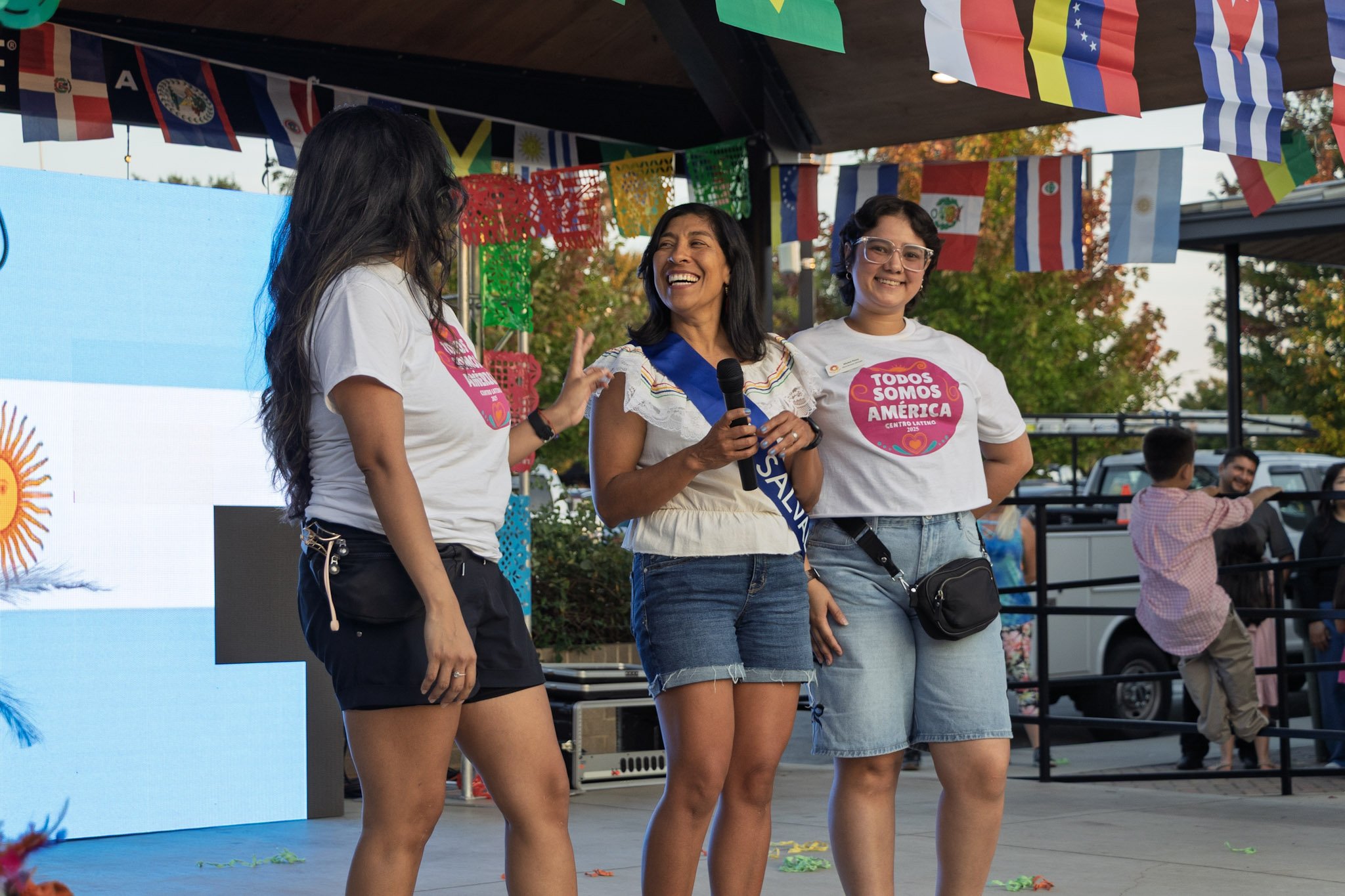 Three women standing on a stage decorated with colorful flags, smiling and talking, with one woman holding a microphone.