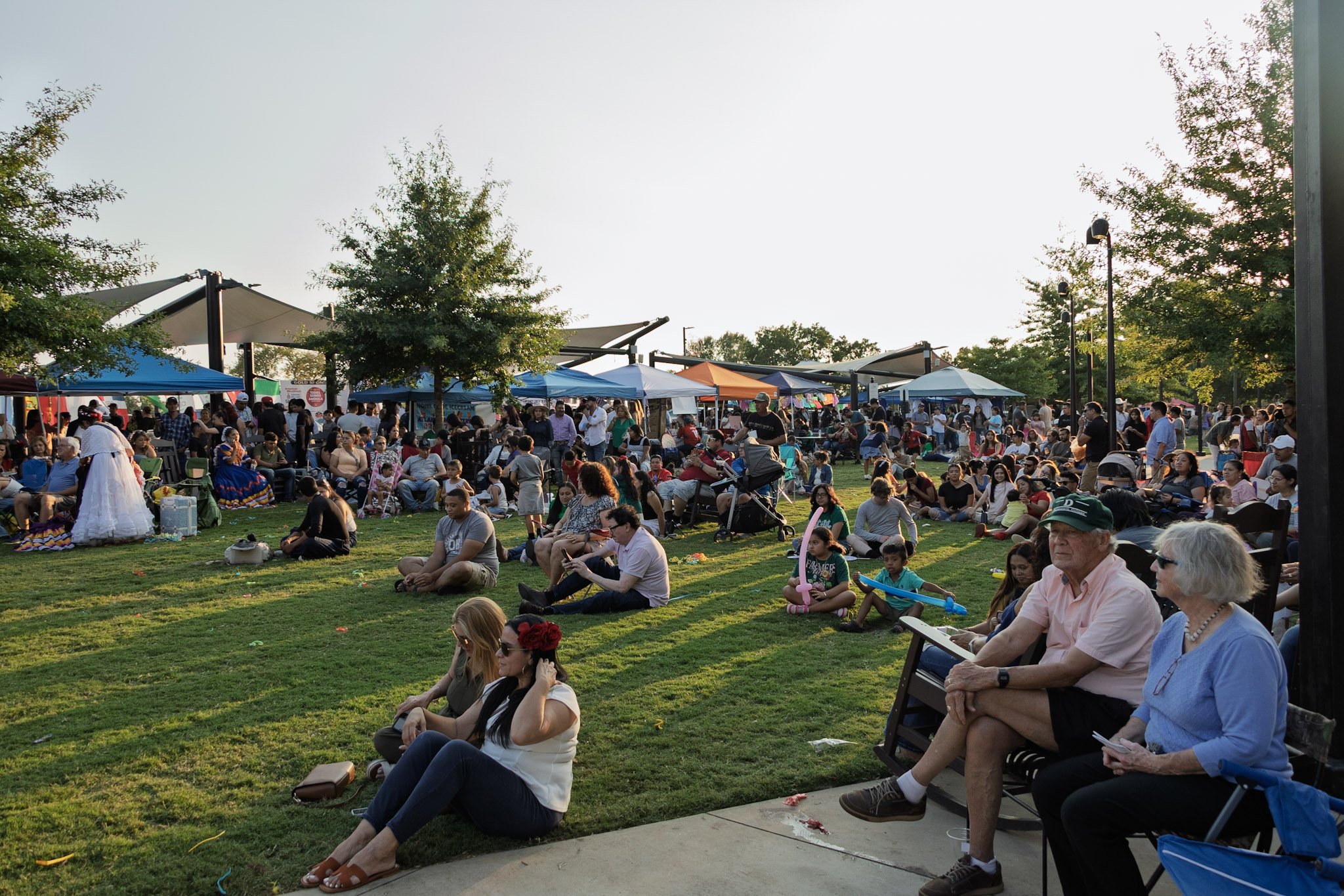 People gathered at an outdoor park event with tents, trees, and a grassy area during late afternoon or early evening.