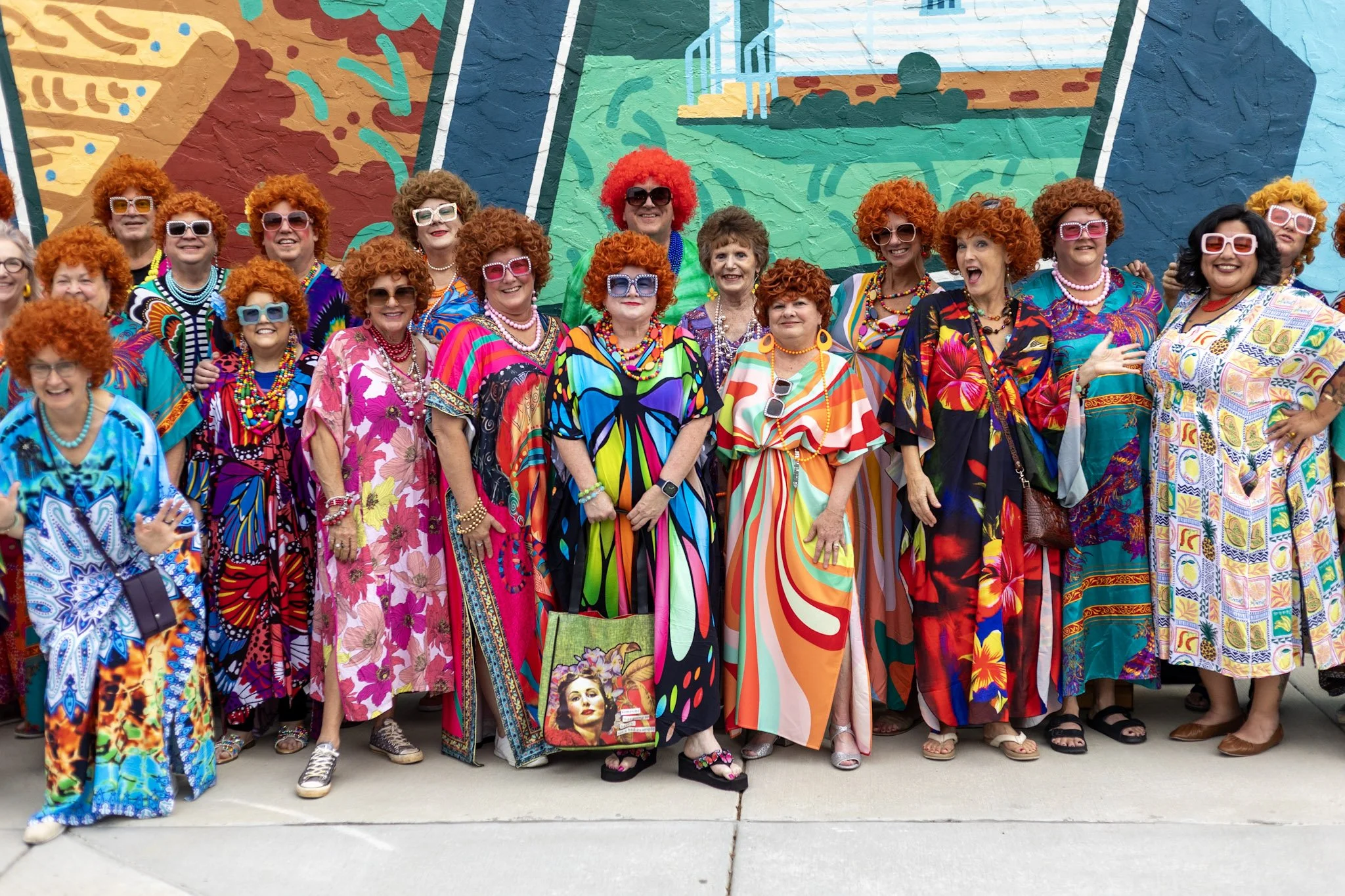 A group of women dressed in colorful, patterned dresses and wearing matching curly red wigs pose in front of a vibrant mural.