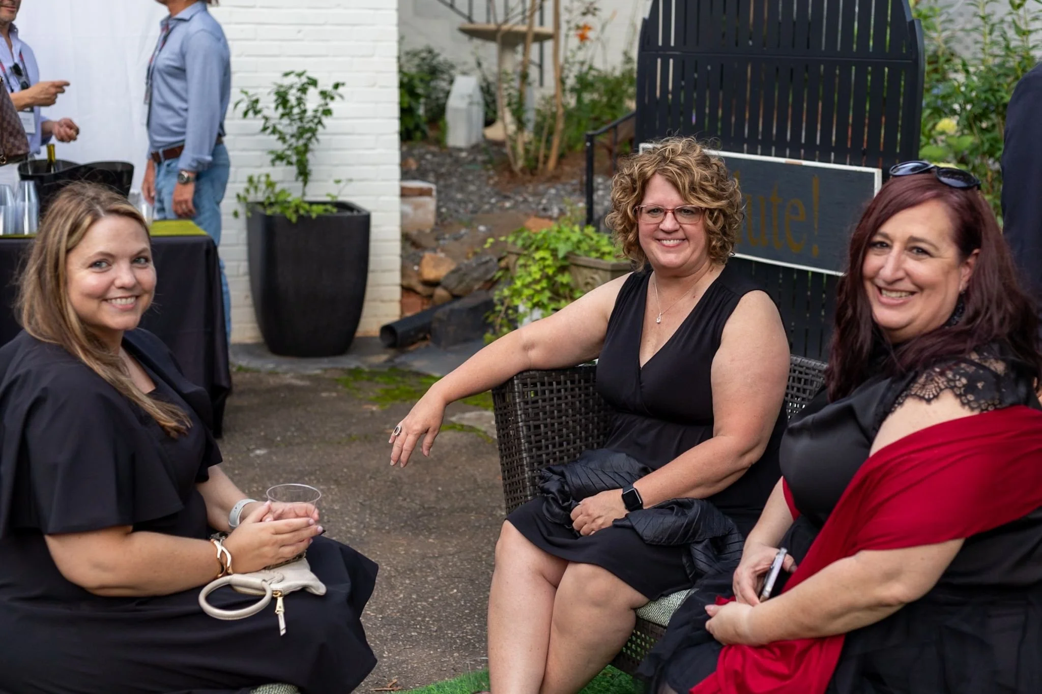Three women sitting outside and smiling, with people in the background, at a social gathering or celebration.