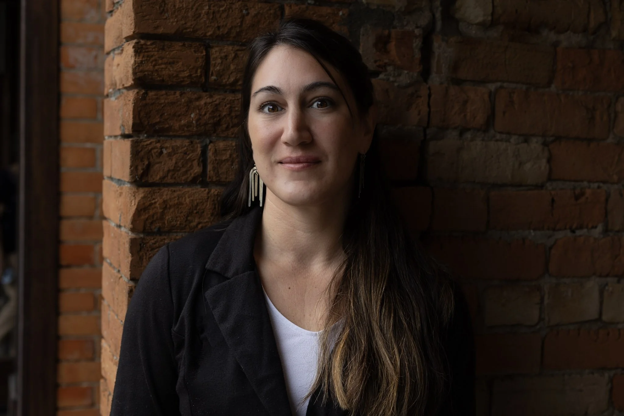 A woman with long brown hair and earrings stands by a brick wall.