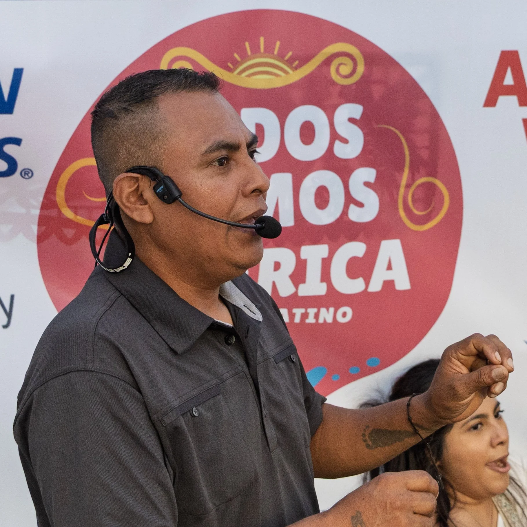 A man wearing a microphone headset speaking, with a woman partially visible behind him, in front of a banner with a red circle and yellow design, and text that reads 'DOS MAMOS AFRICA'.