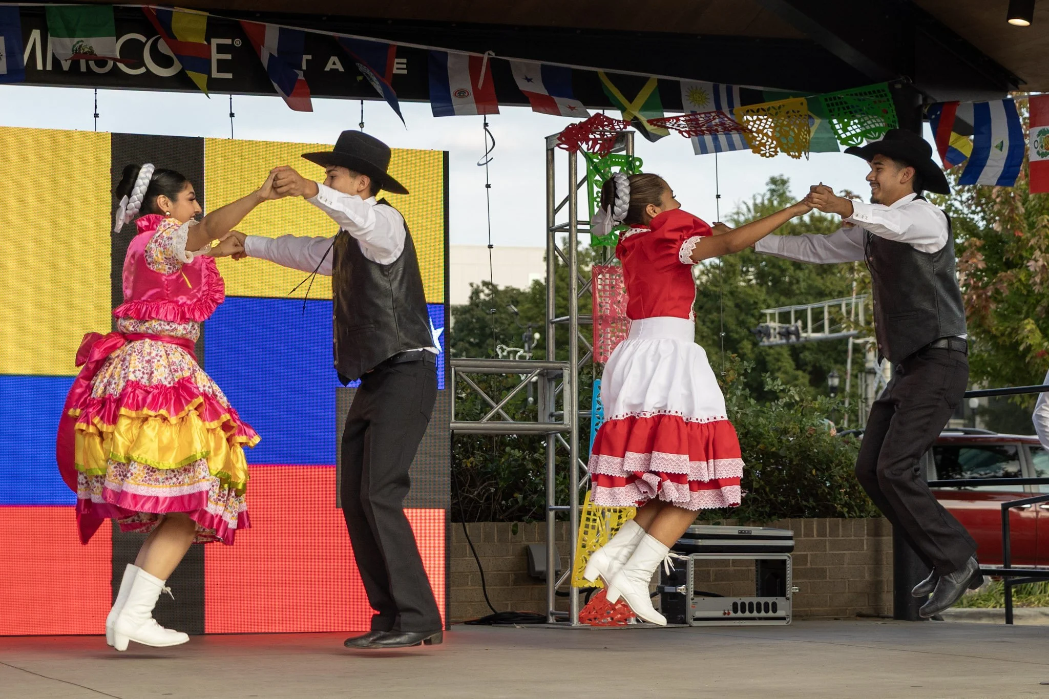 Two couples dancing in traditional Mexican attire on an outdoor stage decorated with colorful banners and flags.