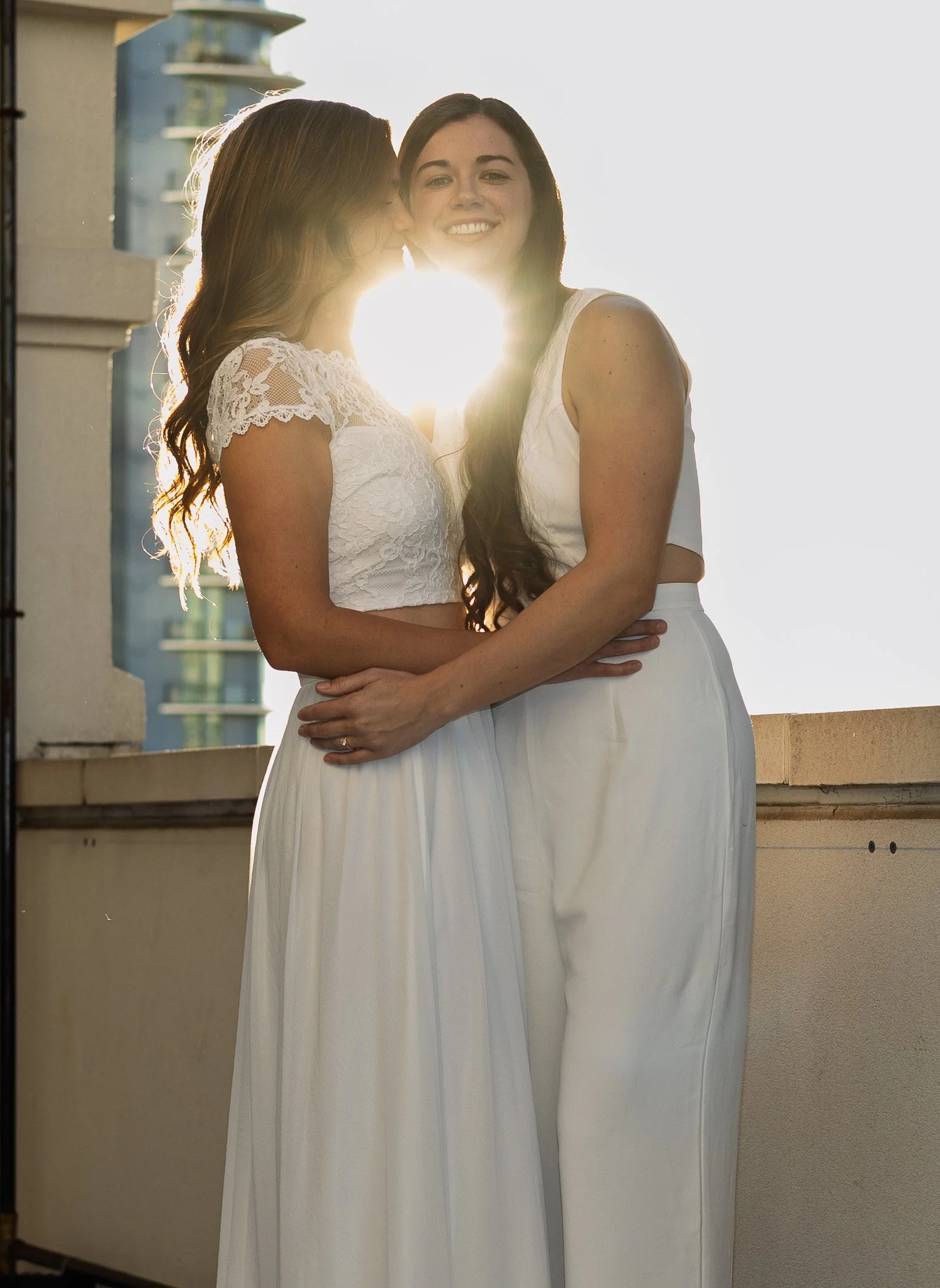 Two women dressed in white, one kissing the other's cheek with the sun setting behind them.