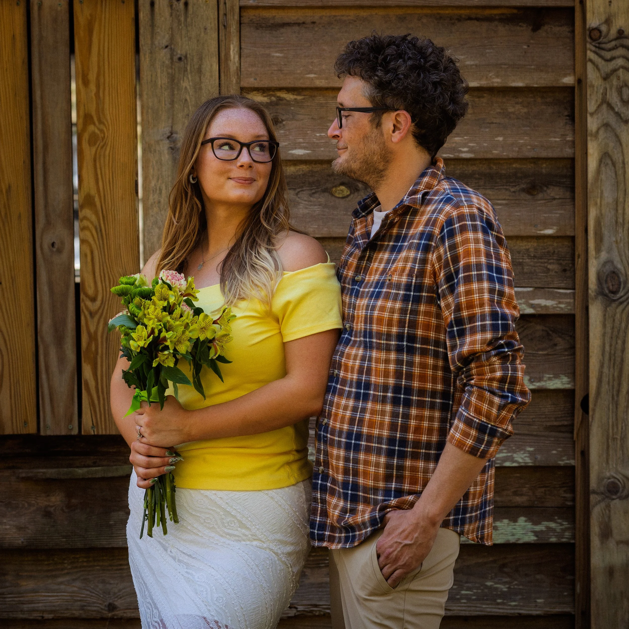 A woman in a yellow top holding a bouquet of flowers stands next to a man in a plaid shirt. They are facing each other and smiling while standing in front of a wooden fence.