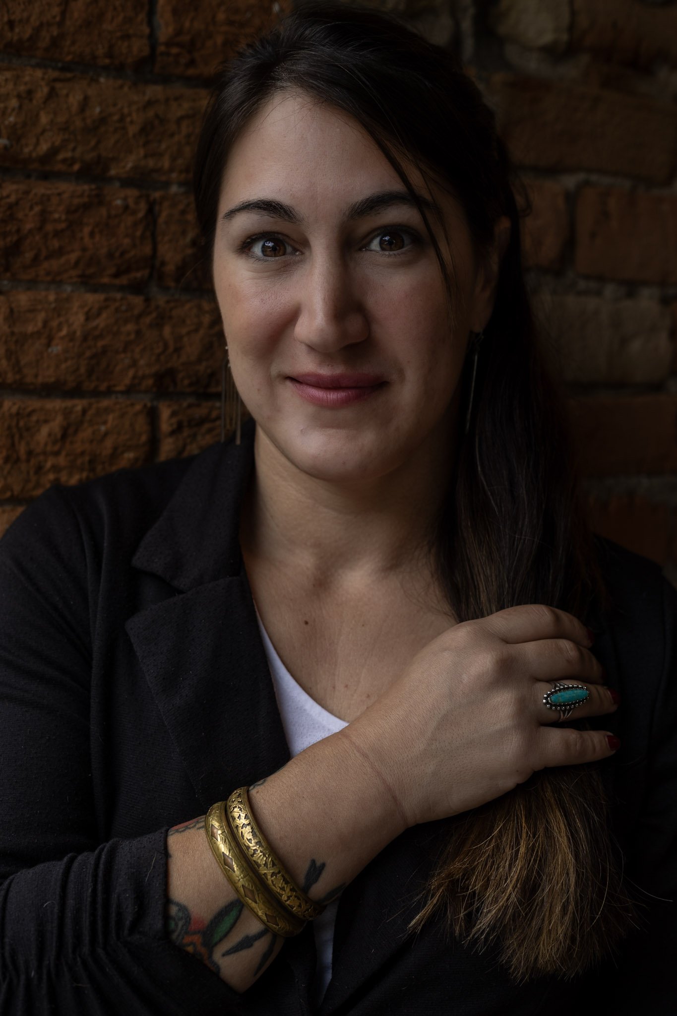 Close-up of a woman with dark hair, wearing a black jacket, gold bracelets, and a turquoise ring, sitting against a brick wall.