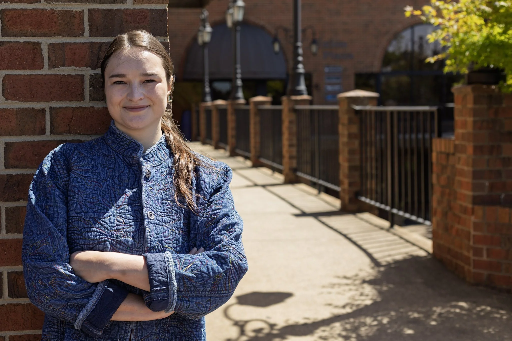Young woman with brown hair in a ponytail, wearing a blue quilted jacket, standing outdoors against a brick wall with arms crossed, smiling slightly, with sunlight and trees in the background.