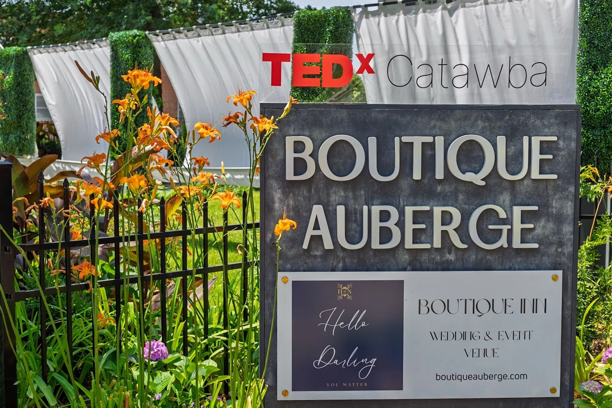 Sign for Boutique Auberge, a wedding and event venue, with a garden of orange and purple flowers in front. The background includes white tents and green foliage, and a large stone sign displays the venue's name.