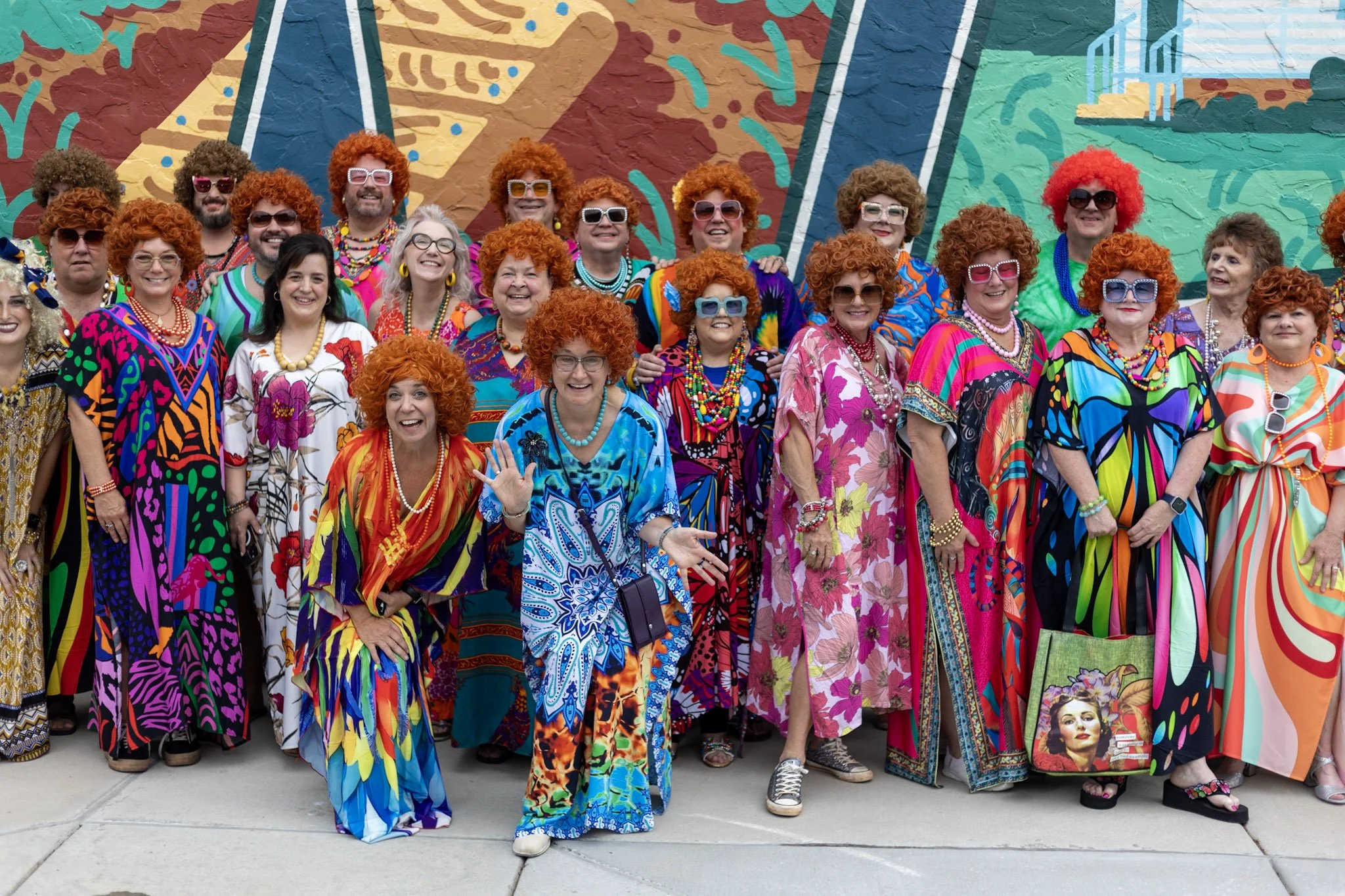 Group of people dressed in colorful, retro outfits with curly red wigs, large sunglasses, and vibrant patterned dresses standing in front of a mural.