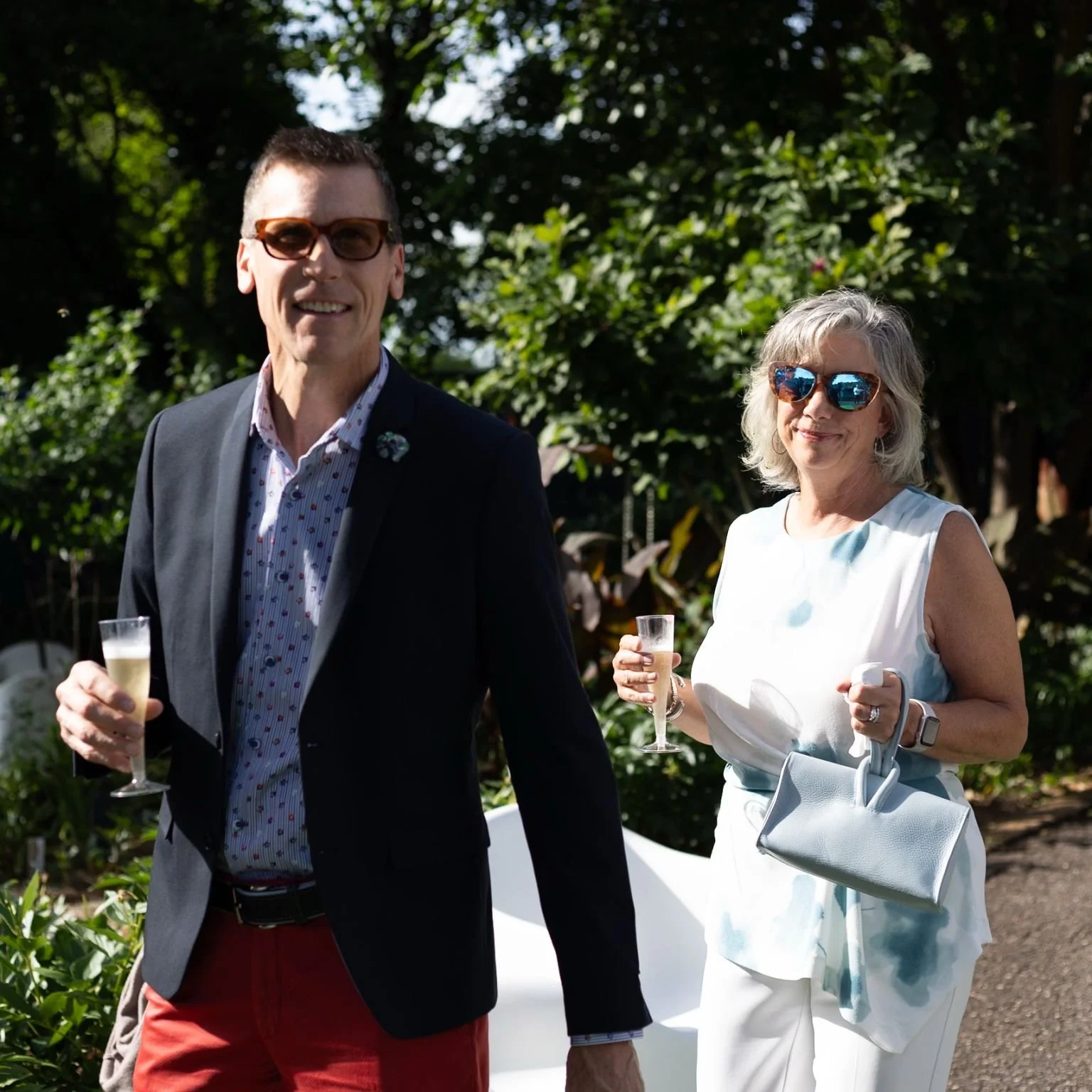A man and woman dressed in semi-formal attire holding glasses of champagne at an outdoor event during daytime. The man is wearing an dark blazer, patterned shirt, red pants, and sunglasses, while the woman is in a white dress, with sunglasses, holdin