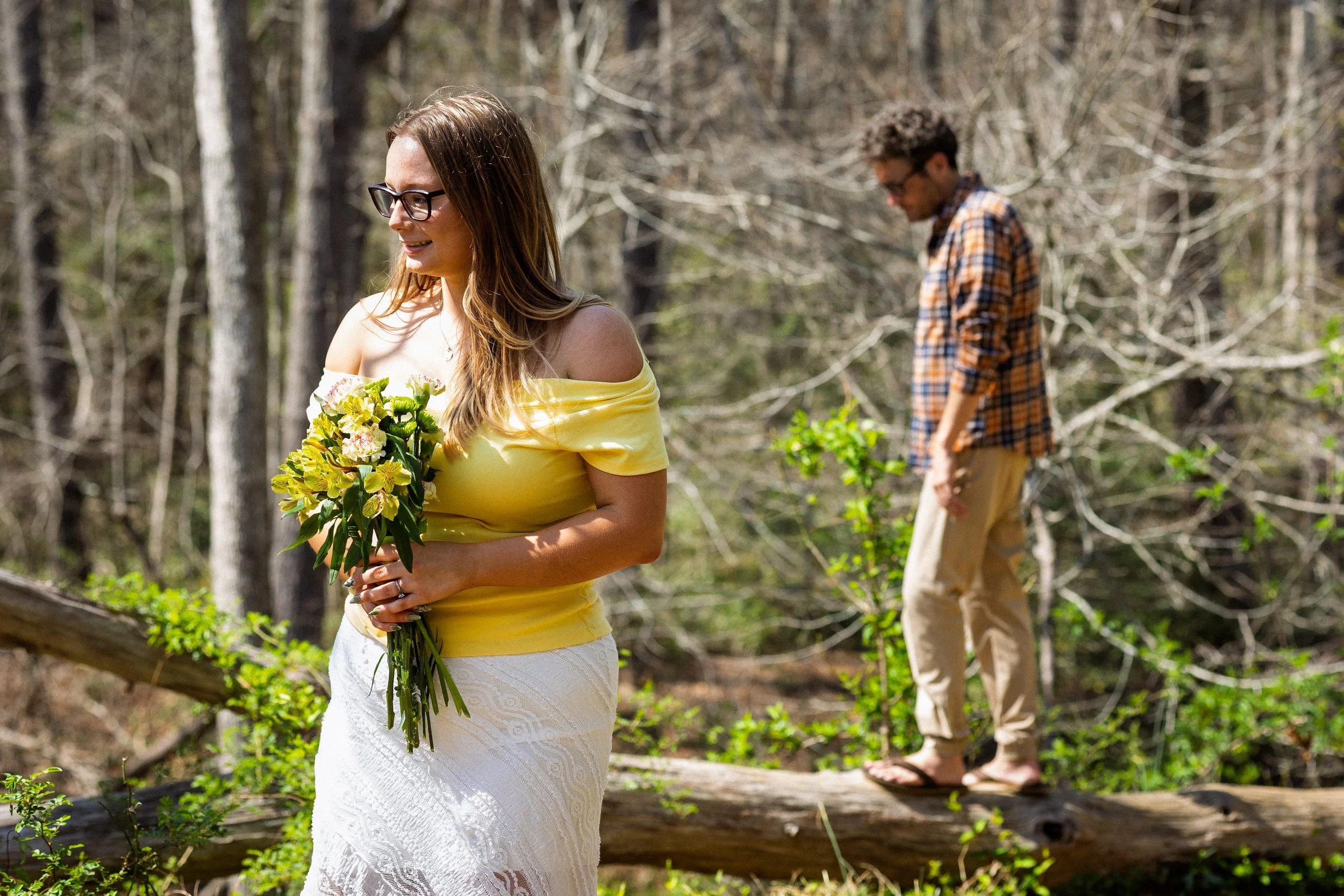 A woman in a yellow top holding a bouquet of flowers, smiling, with a man in a plaid shirt behind her, in a forest setting.