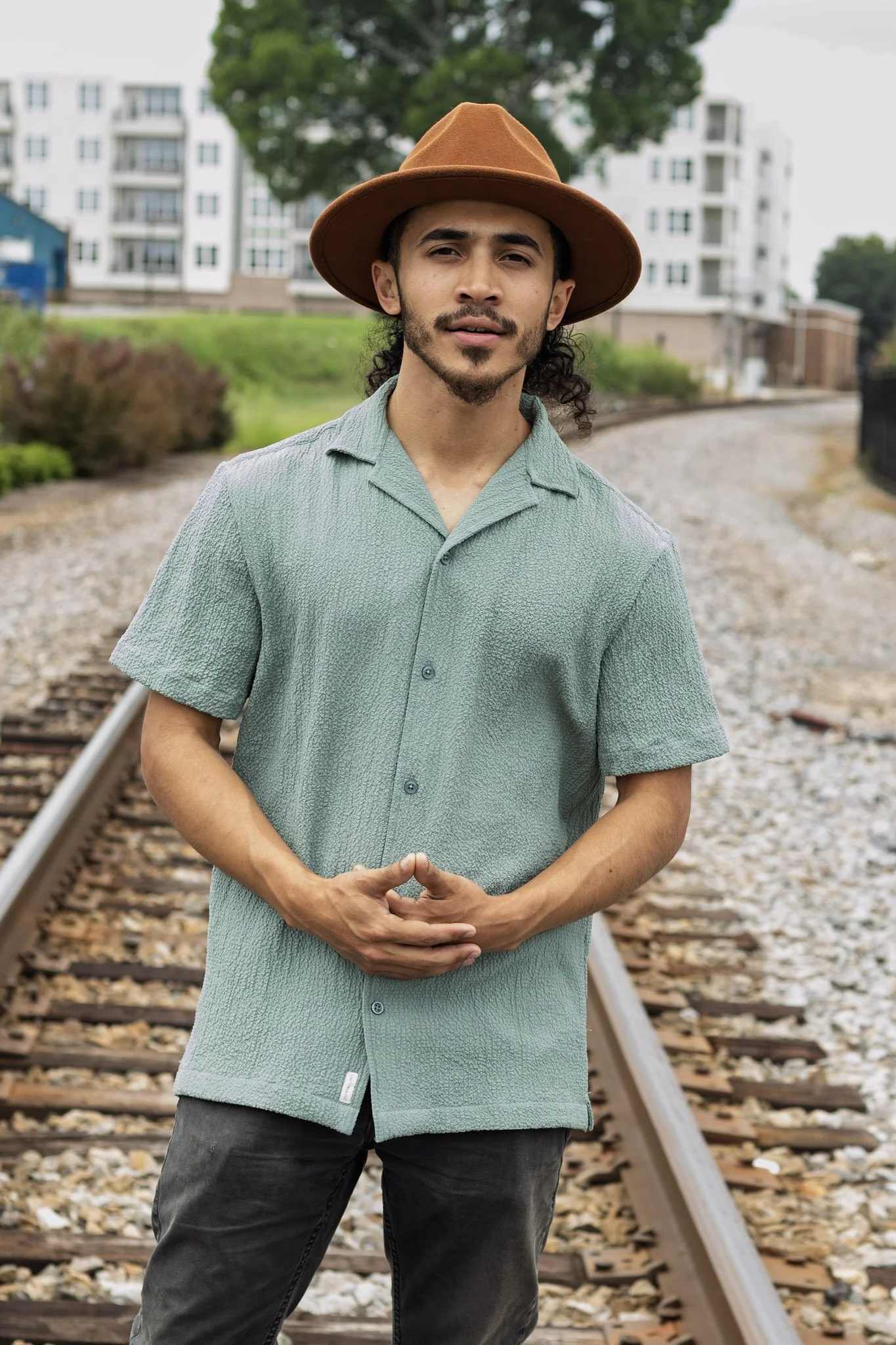 A young man with long curly hair, wearing a brown hat, a short-sleeved light green shirt, and dark pants, standing on a railway track with buildings and greenery in the background.