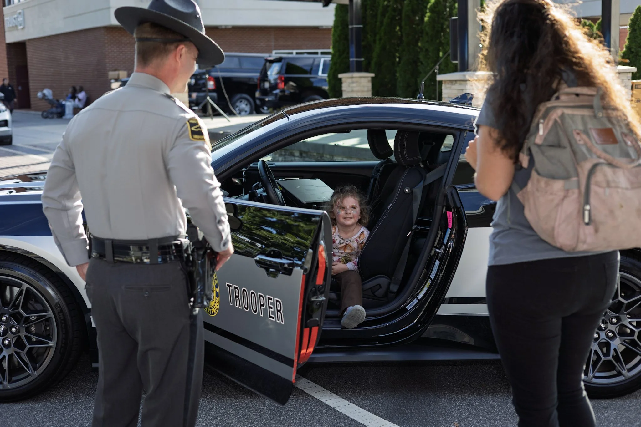 A young girl is sitting in the back seat of a police car with the door open, smiling at two officers who are talking to her. One officer, dressed in a uniform and hat, is standing outside the car, while a woman with curly hair and a backpack is also 
