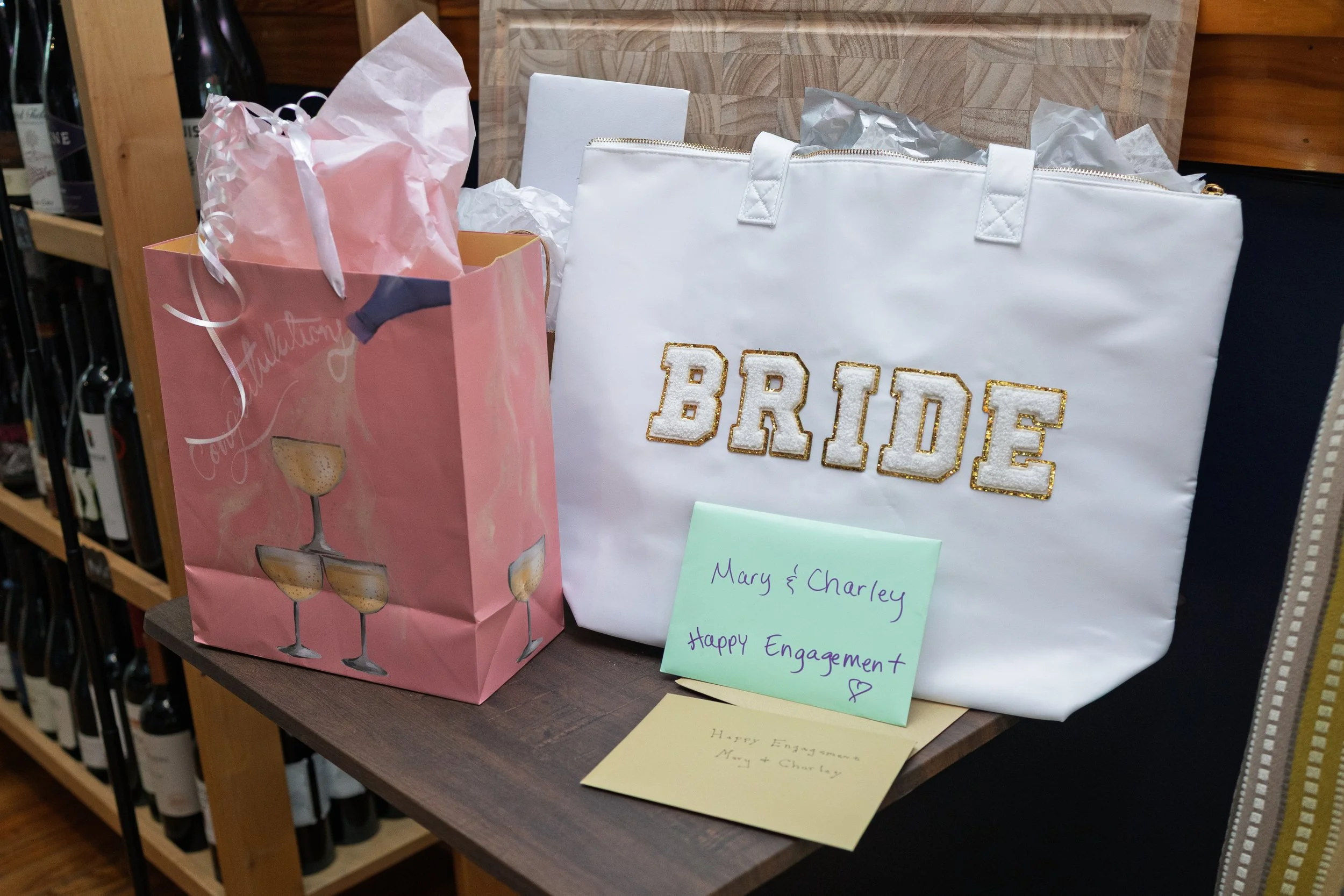The image shows a table with gifts and cards for an engagement celebration. There is a pink gift bag with tissue paper, a white bag with gold decorative lettering spelling 'BRIDE,' and handwritten notes that read 'Mary & Charley Happy Engagement' and