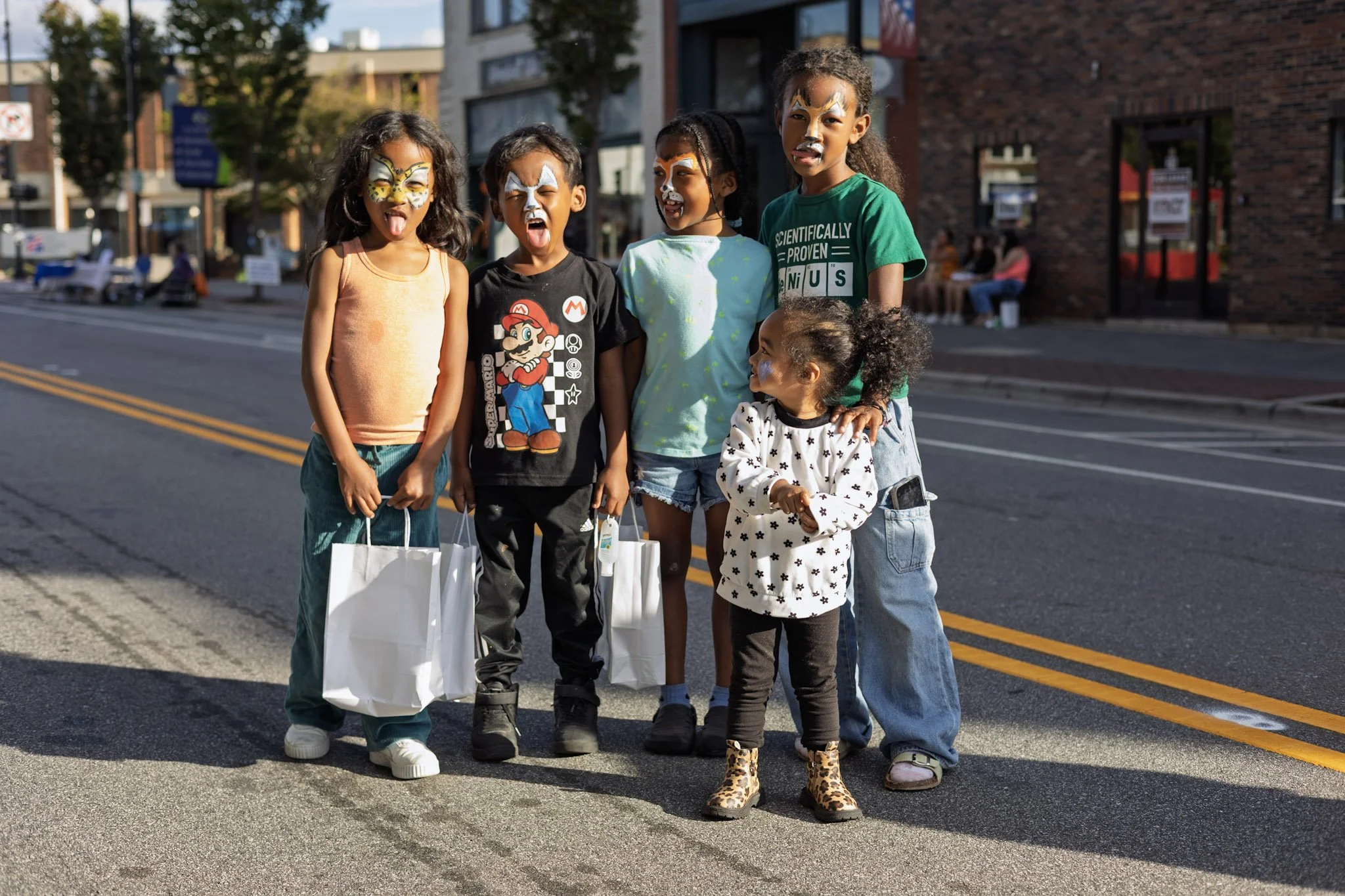 Group of six children with face paint, standing on street with shopping bags, smiling and making silly faces.