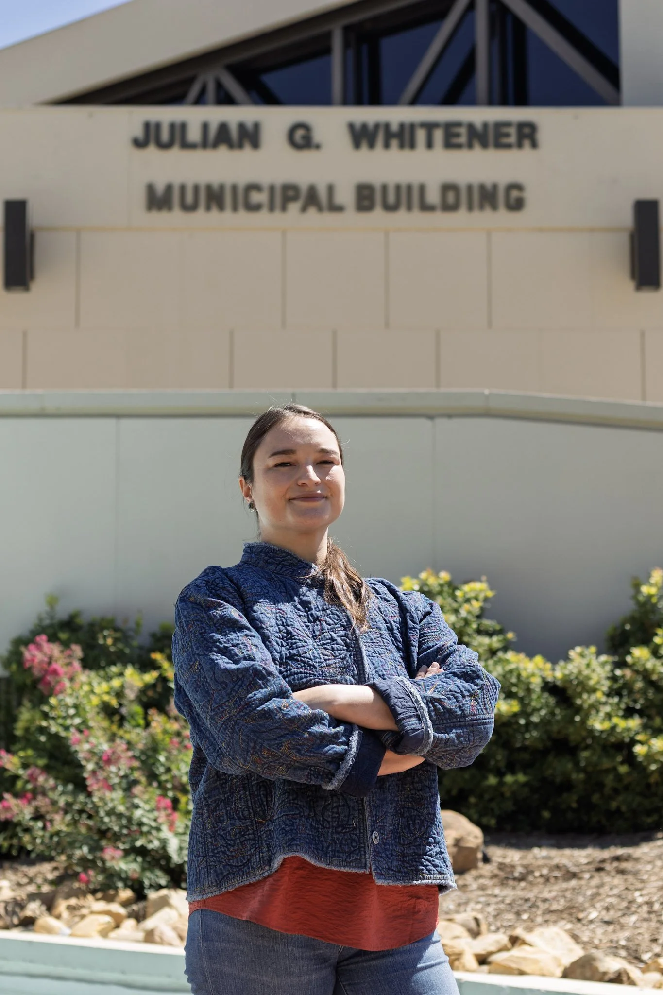 Woman standing outside in front of the Julian G. Whitener Municipal Building with arms crossed, smiling, with shrubs and flowers behind her.