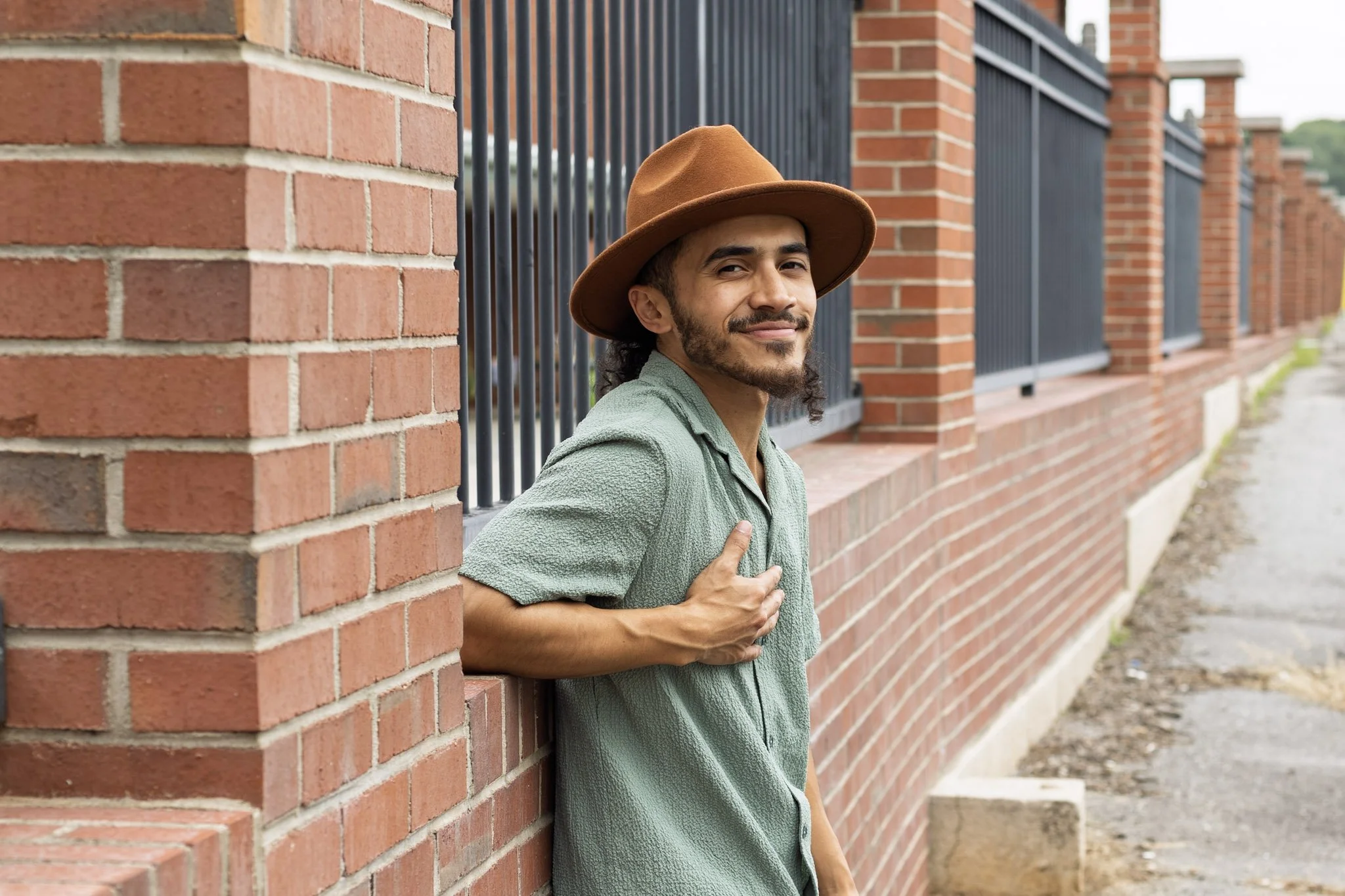 A man leaning against a brick wall, wearing a brown hat and a green shirt, smiling at the camera.