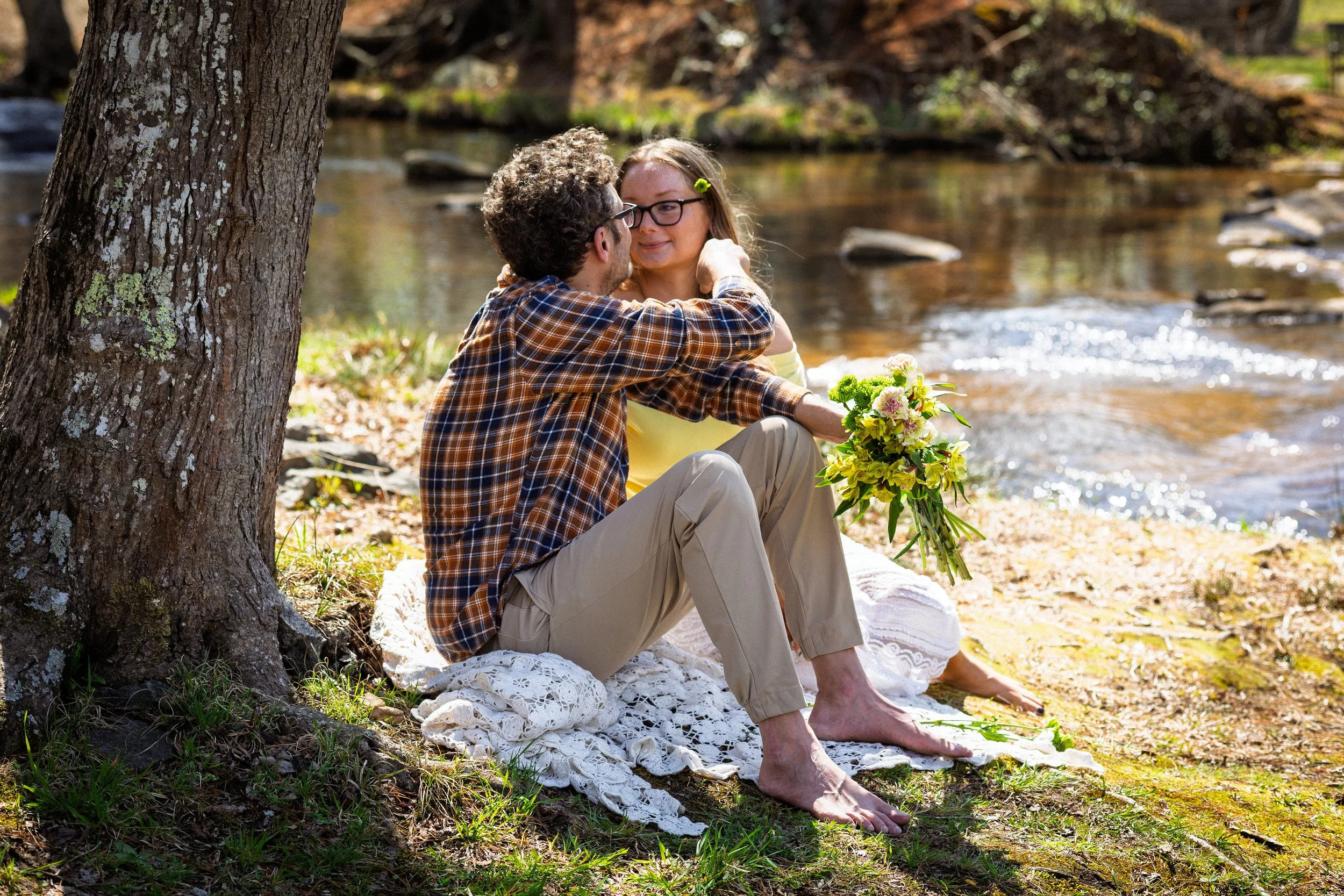 A couple sitting by a river on a sunny day, with the woman holding a bouquet of yellow flowers. The woman is wearing glasses and a yellow top, while the man is wearing a plaid shirt and beige pants. They are sitting on a white lace blanket, barefoot,